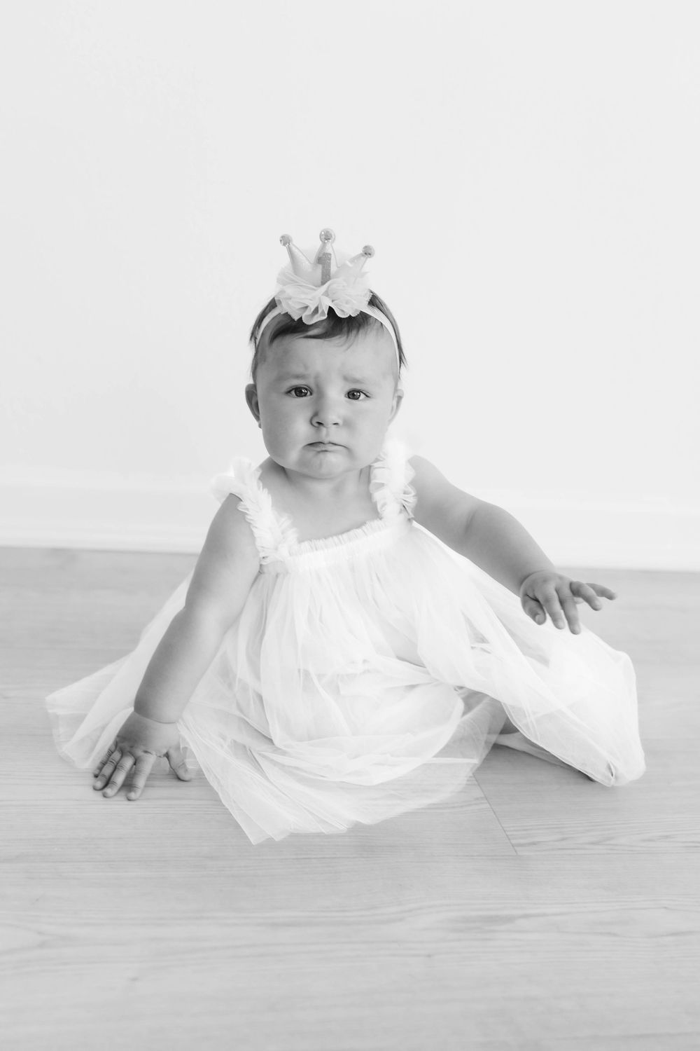 Black and white portrait of a baby girl in a white dress and crown headband sitting on the floor.