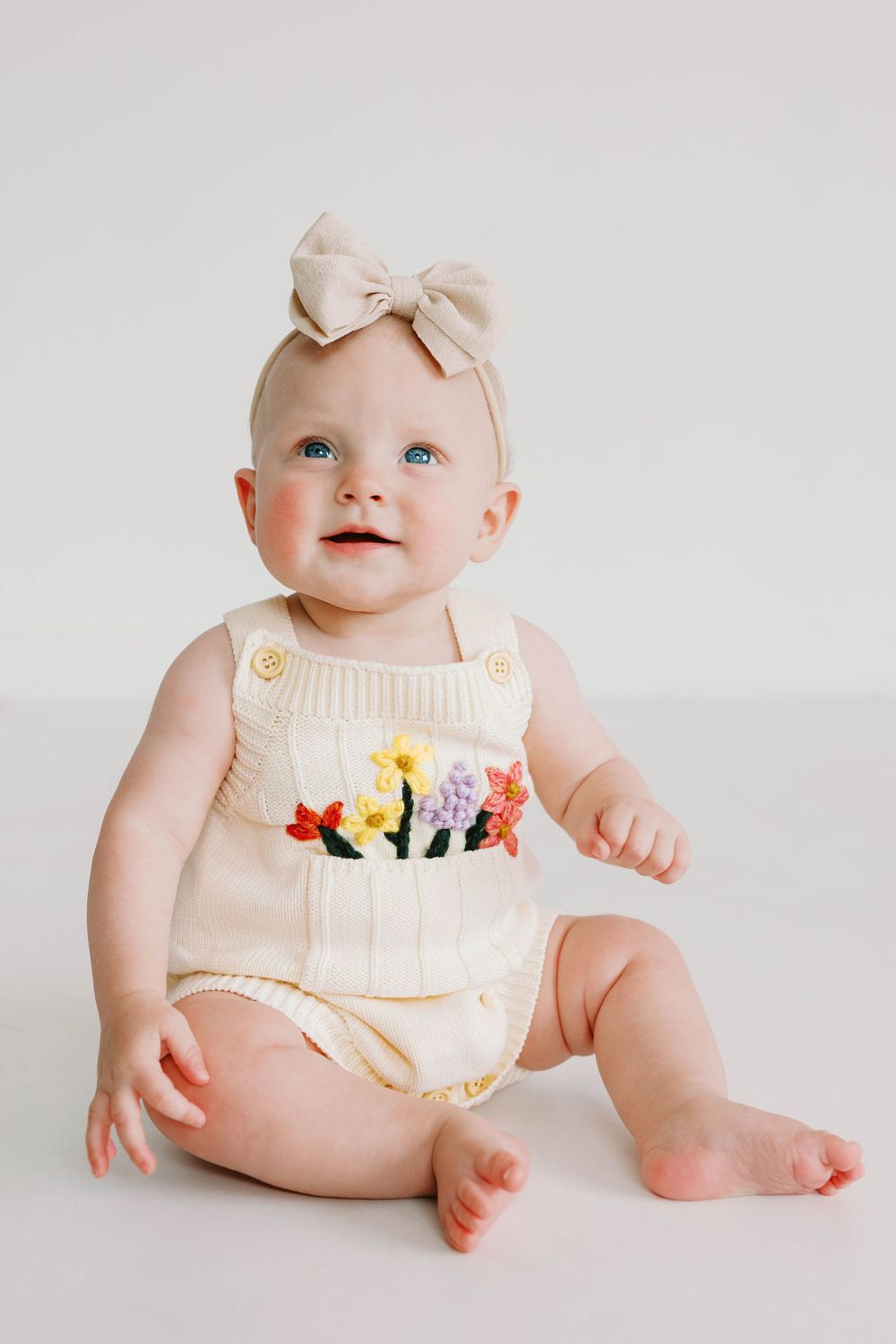 Baby girl with blue eyes sitting in a floral knit romper and beige bow headband.