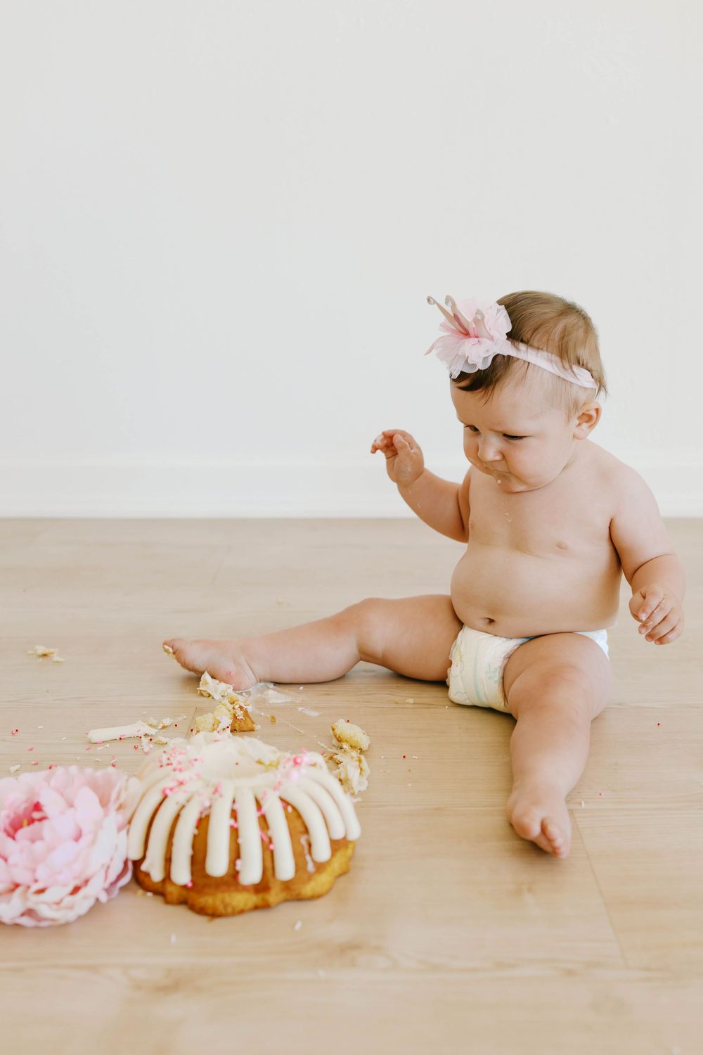 Baby girl in a diaper and floral headband sitting on the floor next to a smashed birthday cake.