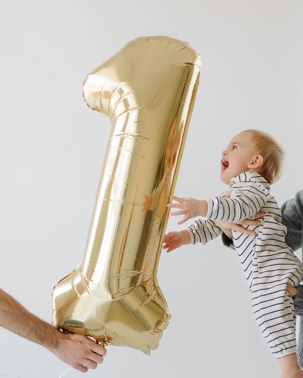 Excited baby in a striped onesie reaching for a large gold number one balloon.