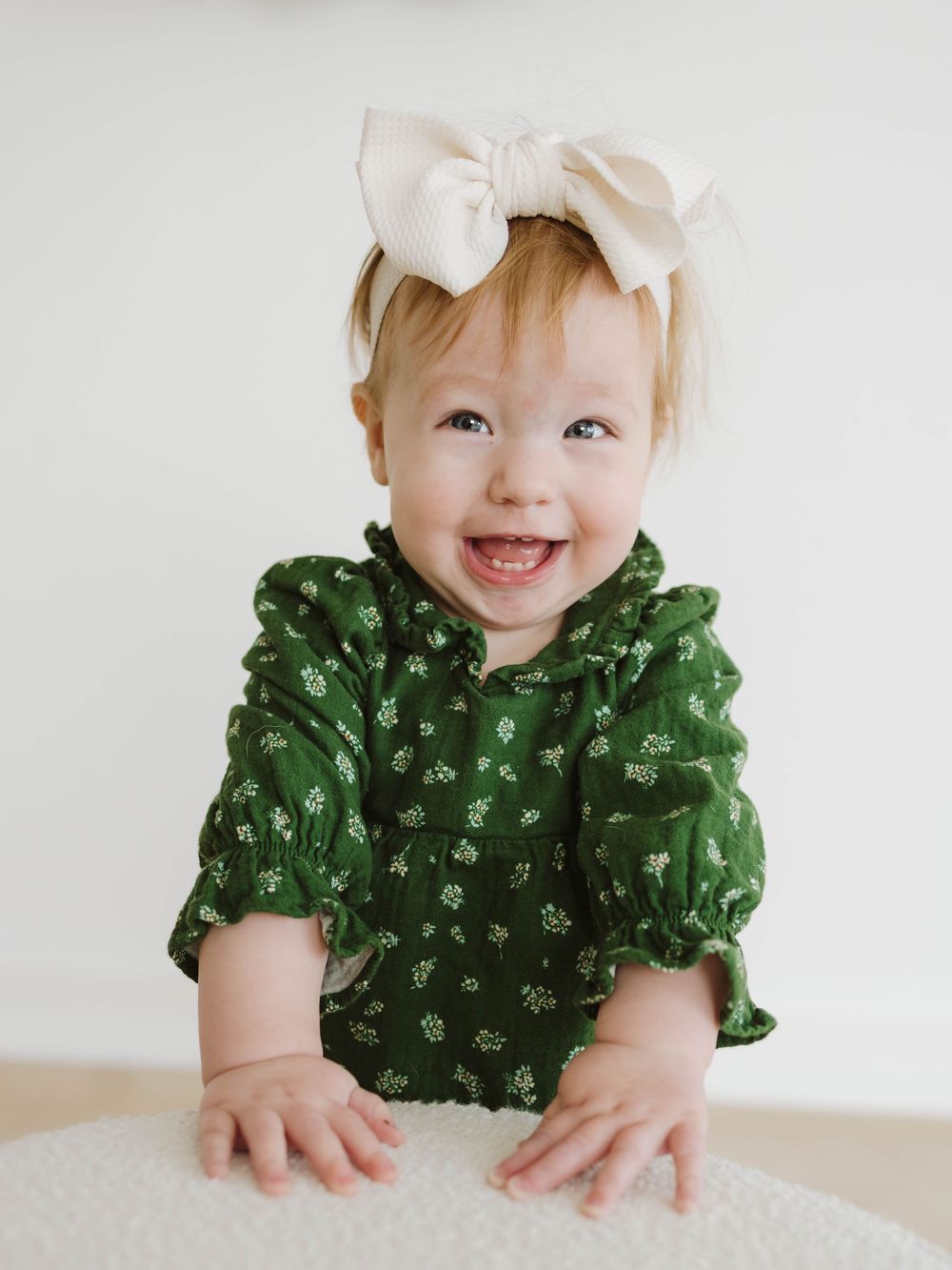 Cheerful baby girl in a green floral romper and cream bow headband smiling brightly.