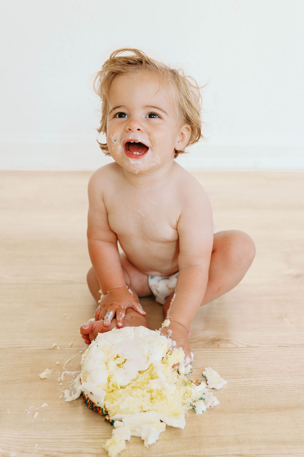 Happy toddler in a diaper smashing a white frosted cake on a wooden floor.