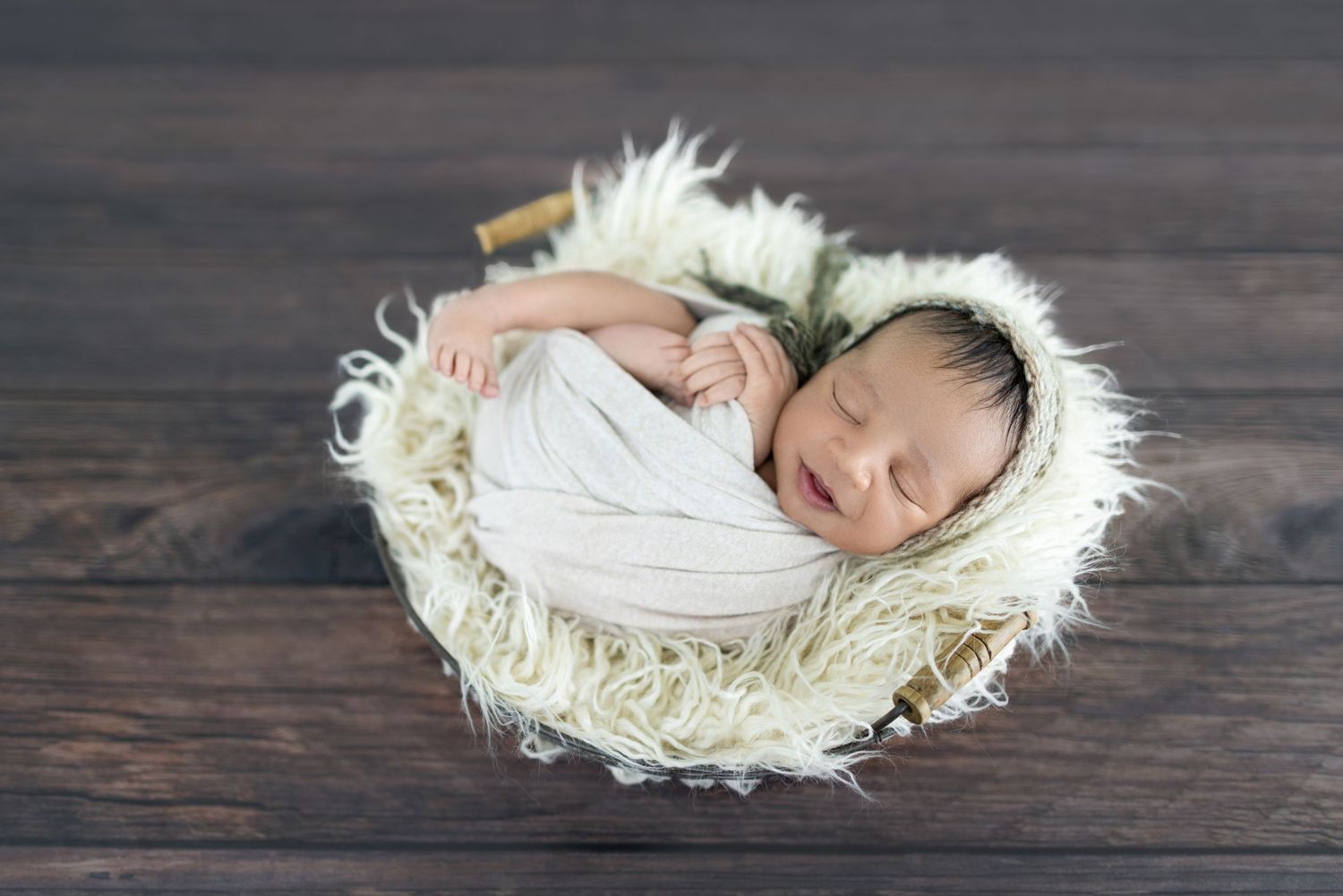 Newborn baby swaddled in white wrap sleeping peacefully in a rustic basket lined with fluffy cream fur.