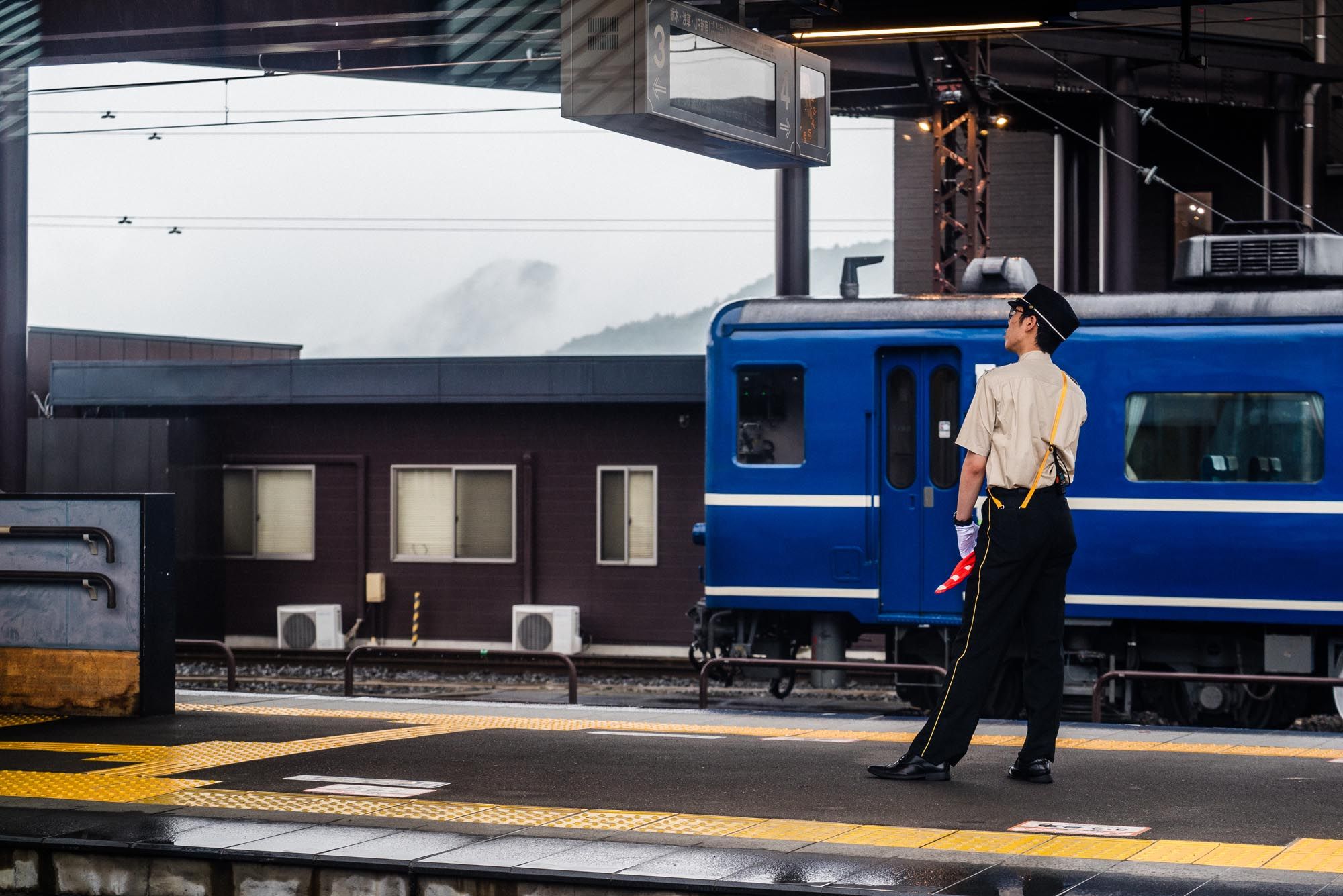 A Japanese railway conductor stands on a train platform beside a blue passenger train at a station.
