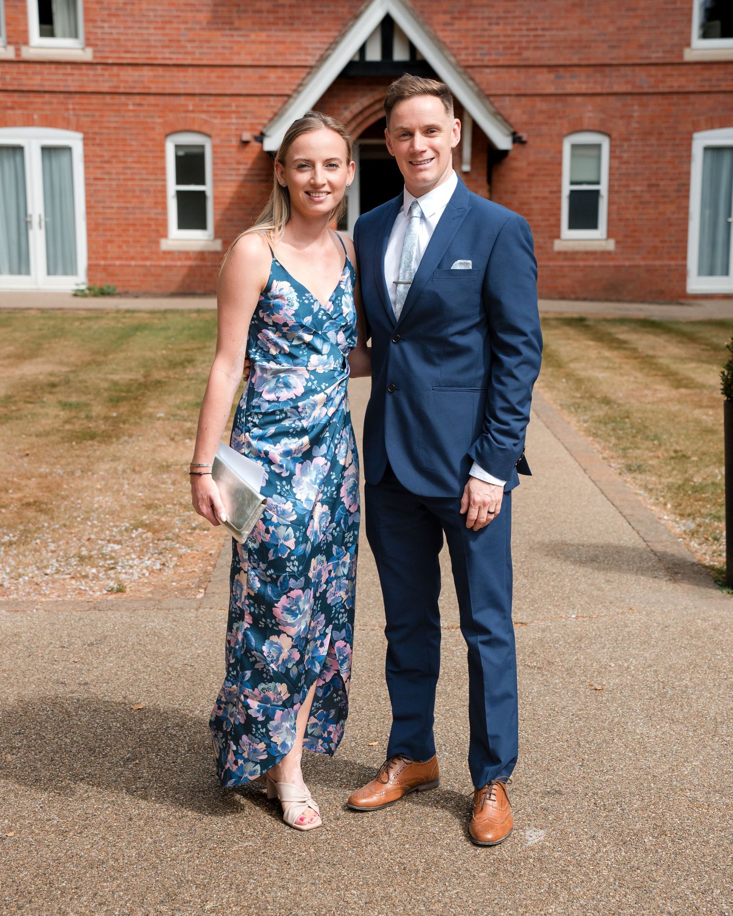 A couple poses outdoors in formal attire, woman in floral dress, man in navy suit, before a red brick building.