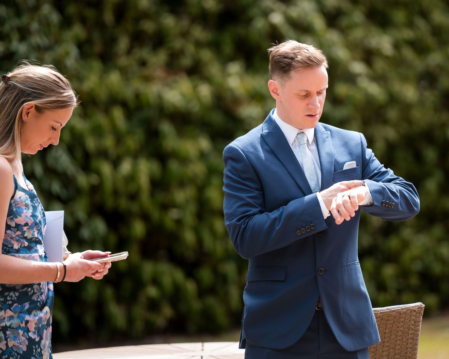 Man in navy suit checks watch outdoors while woman in floral dress reads from phone at a garden event.