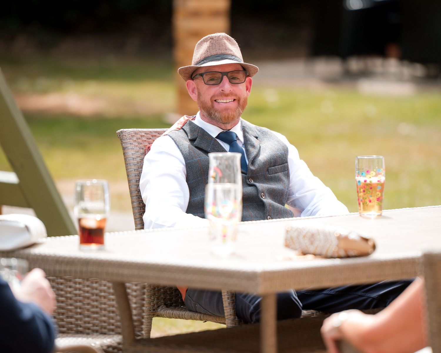Smiling bearded man in a gray vest, blue tie, and brown fedora sitting at an outdoor table with drinks.
