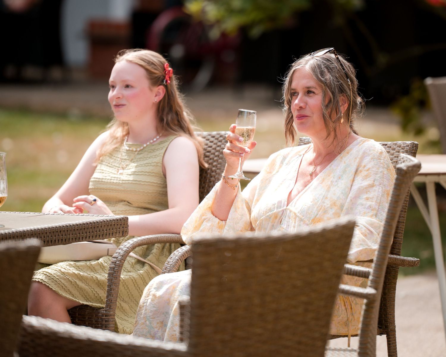 Two women in elegant dresses sit outdoors holding champagne glasses at a sunny garden event.