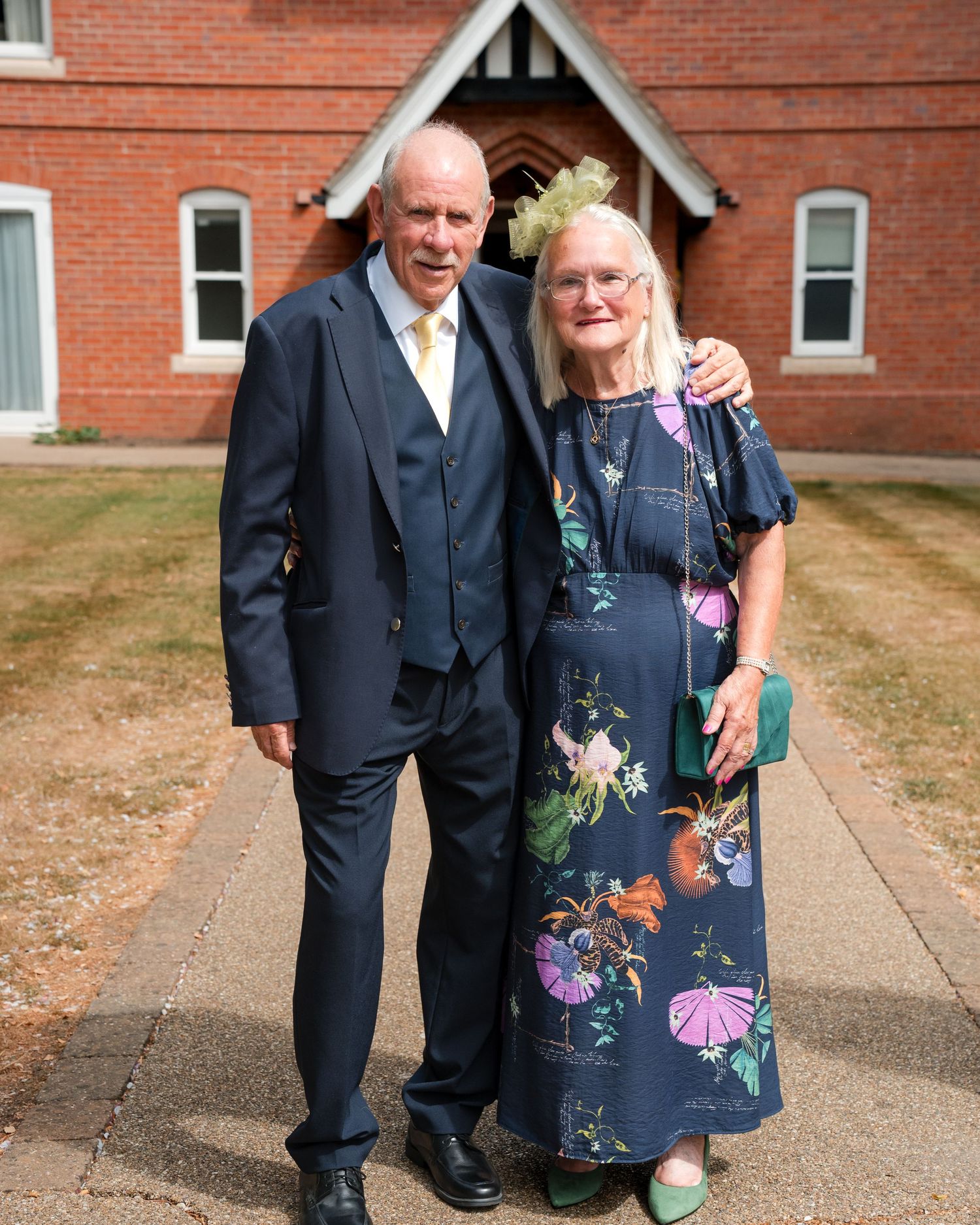 Elderly couple posing together outside a brick building, man in navy suit, woman in floral dress with green fascinator.