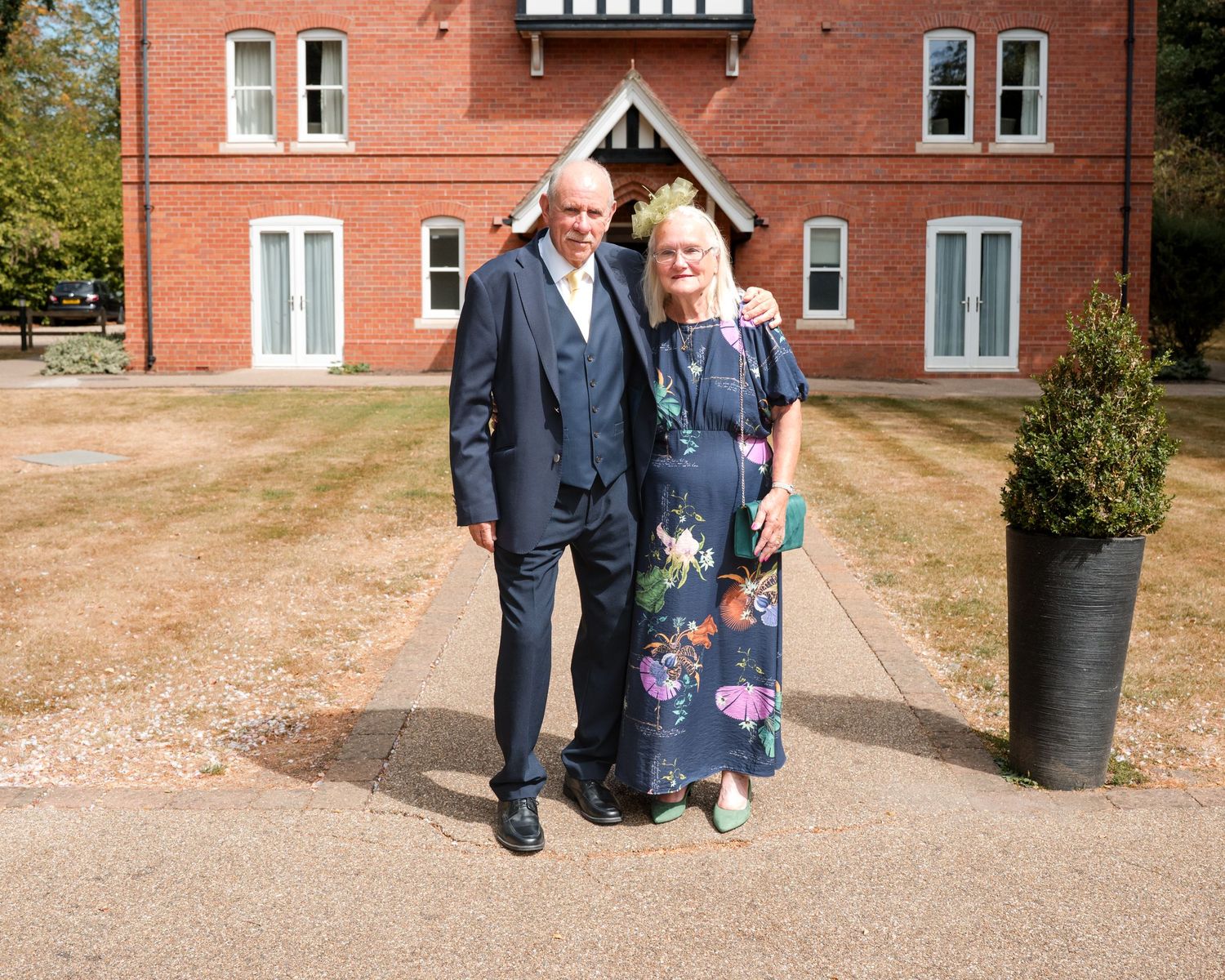 Elderly couple posing together outside a red brick building on a sunny day, dressed in formal attire.