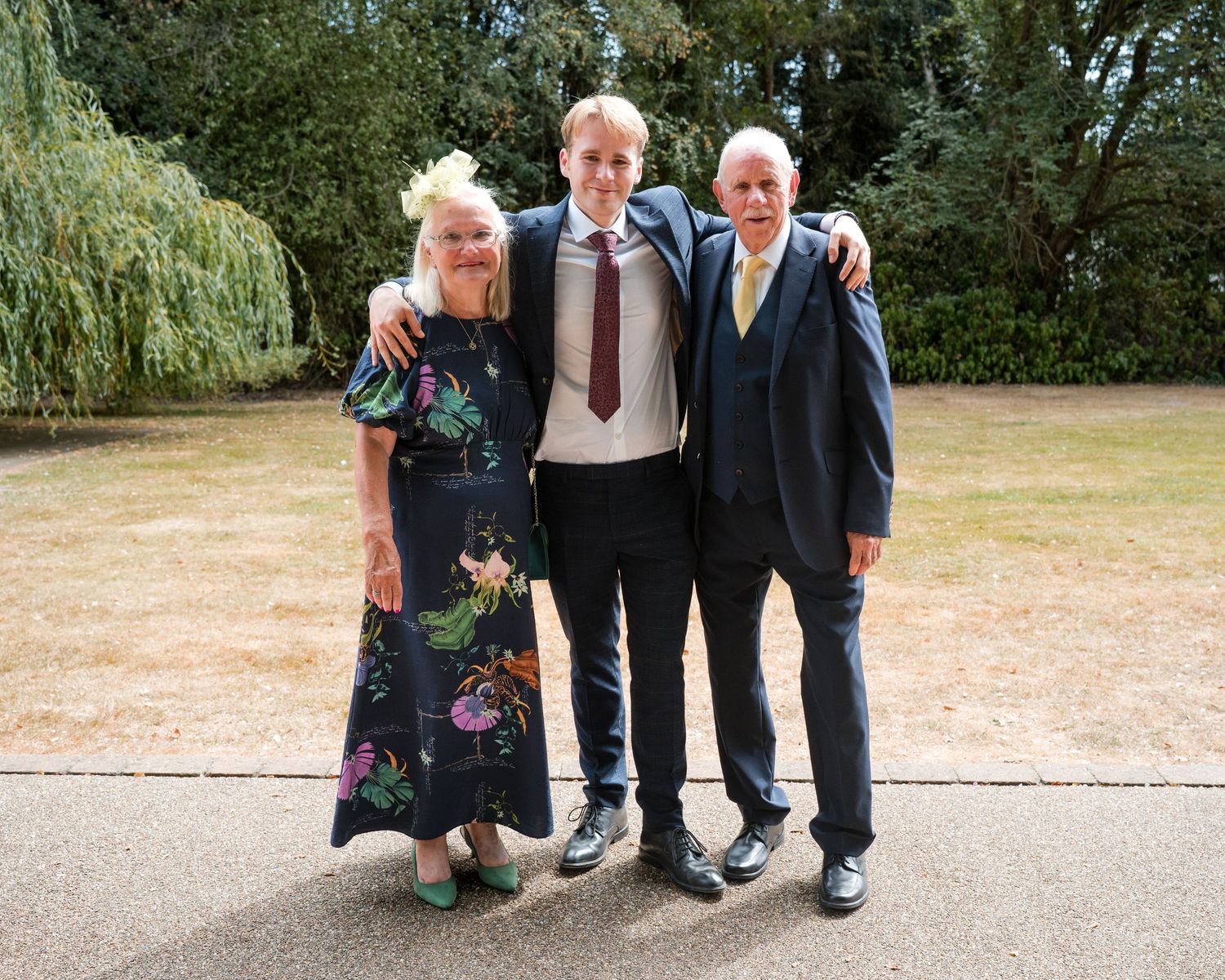 Three people posing together outdoors at a formal event, with lush green trees in the background.