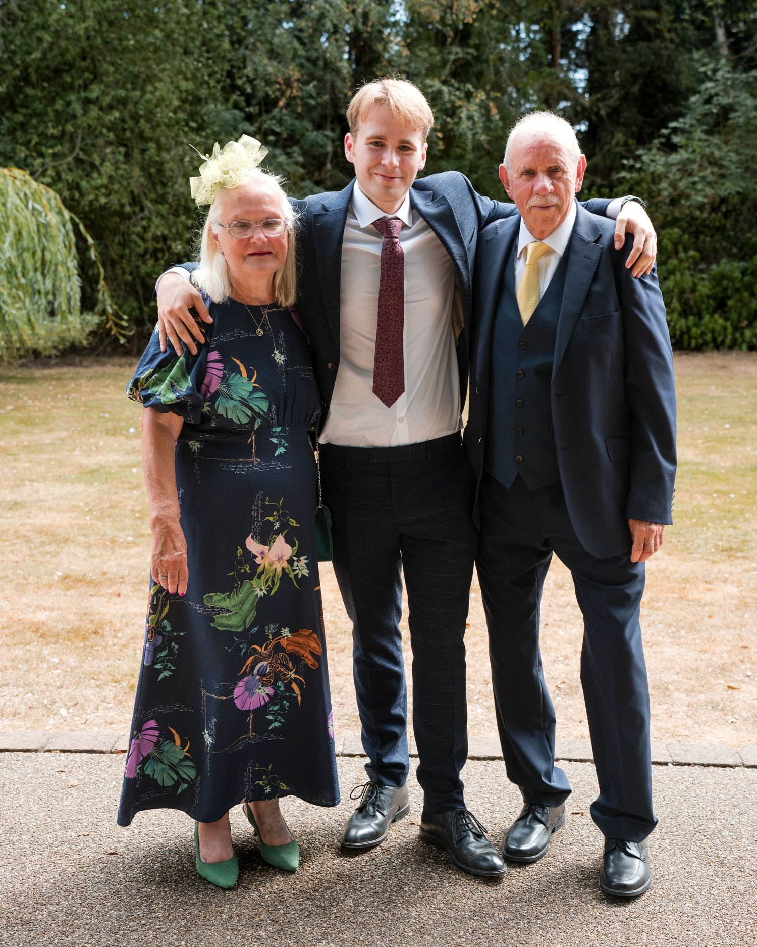 Three people posing together outdoors at a formal event, woman in floral dress, tall young man in suit, older man in navy suit.