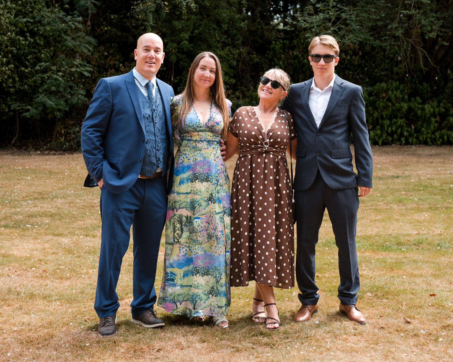 Four elegantly dressed people posing together outdoors on grass at a formal garden event.
