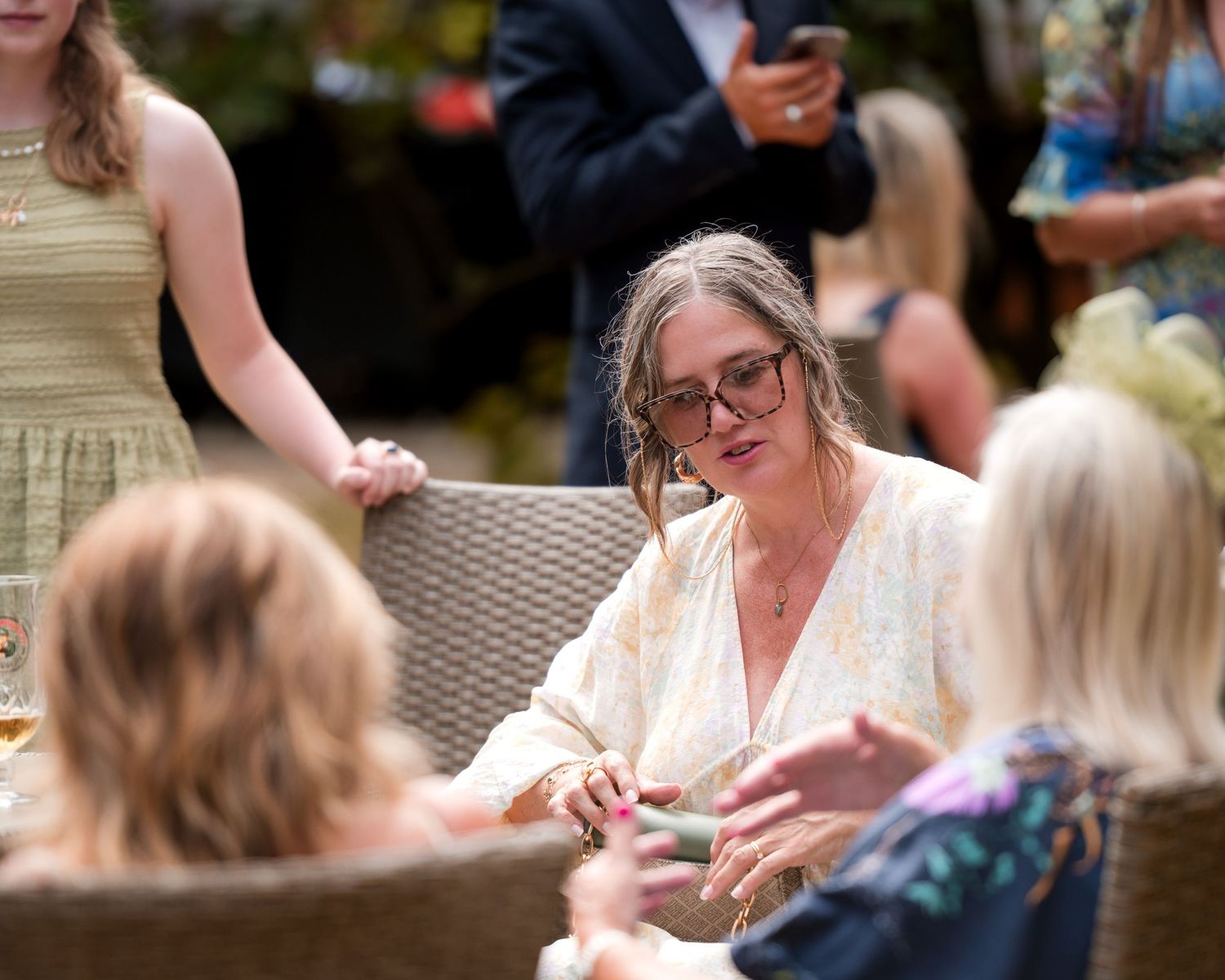 Woman with glasses in floral dress sits at outdoor garden party, engaged in conversation with guests.
