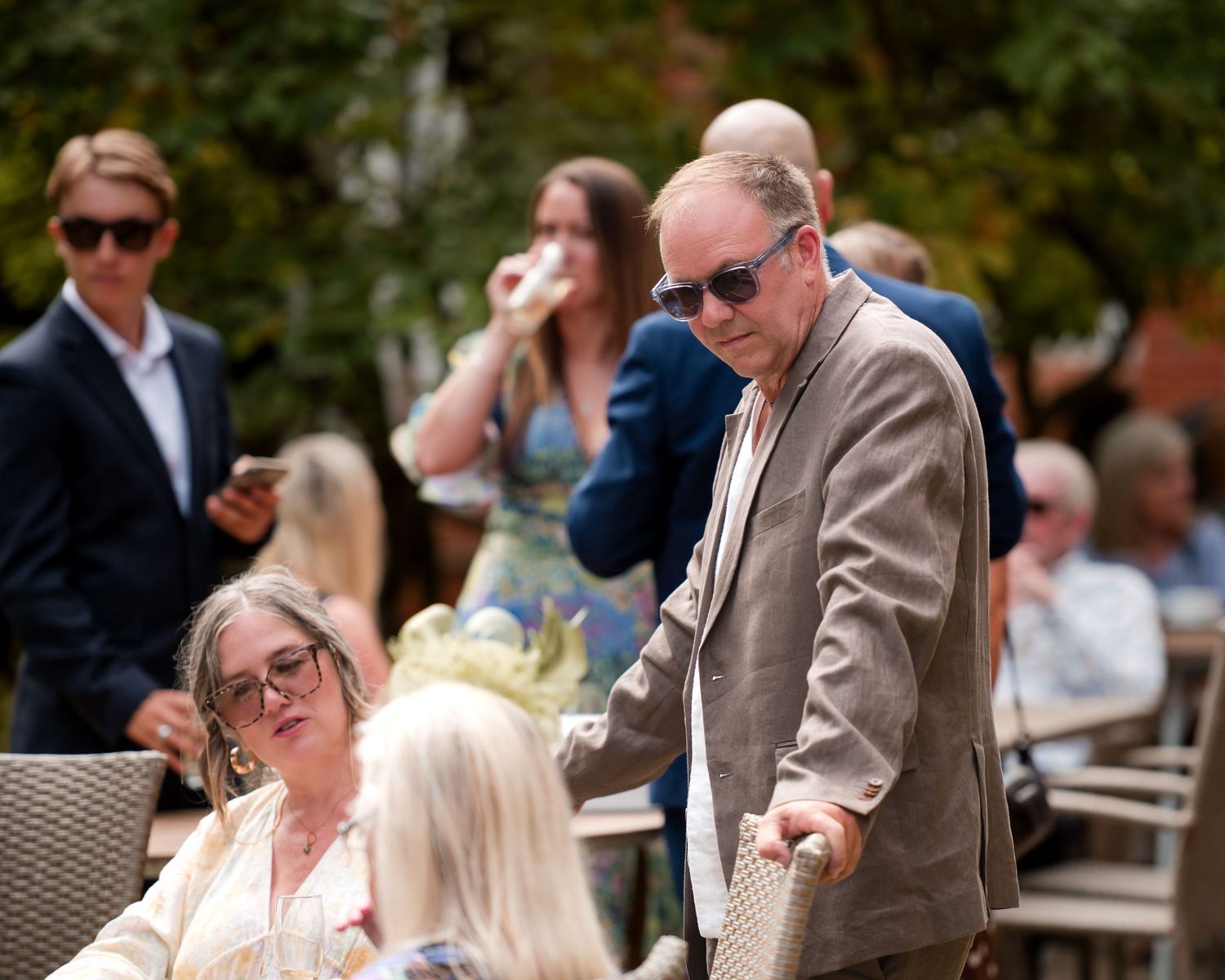 Elderly man in sunglasses and beige blazer greeting seated guests at an outdoor garden party event.