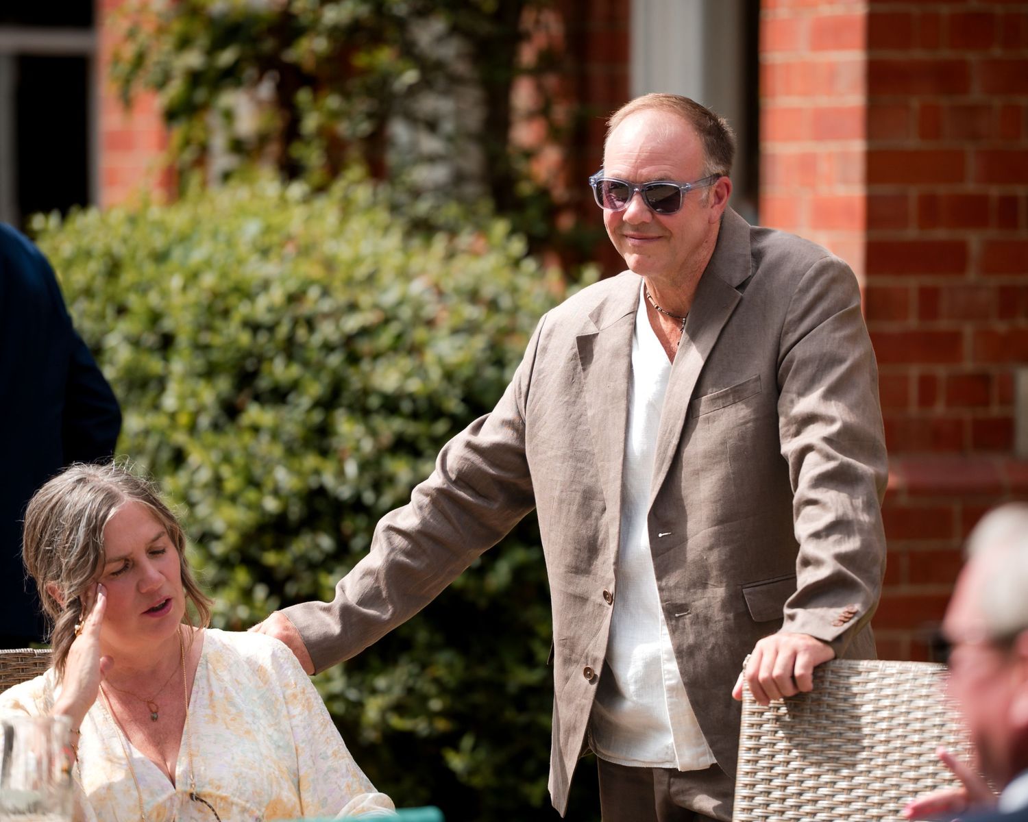 An older man in sunglasses and a grey blazer stands outdoors near a brick building beside a seated woman.