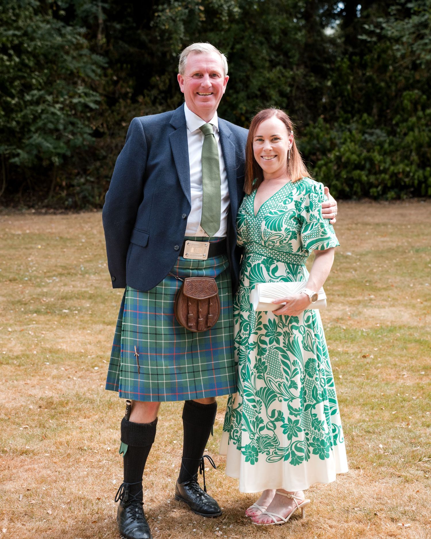 Couple posing outdoors, man in green tartan kilt and blazer, woman in green floral dress, at a formal event.