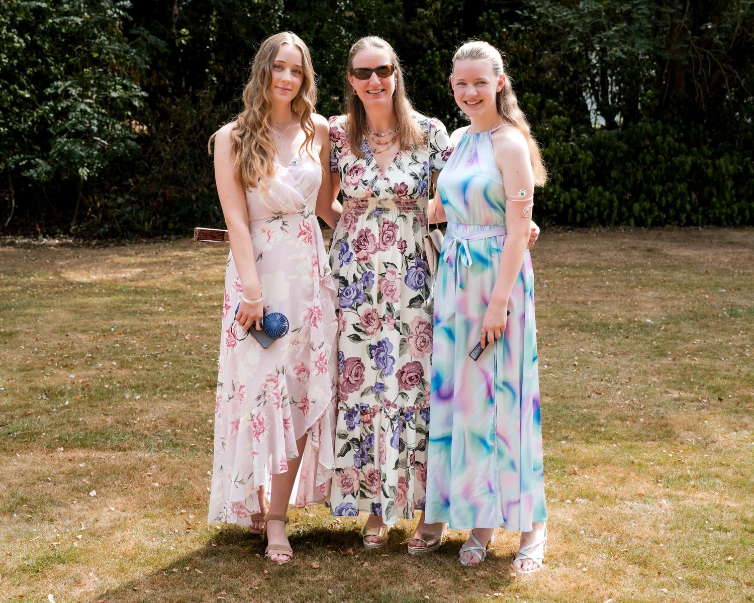 Three women in floral maxi dresses posing together outdoors on a sunny day in a garden setting.