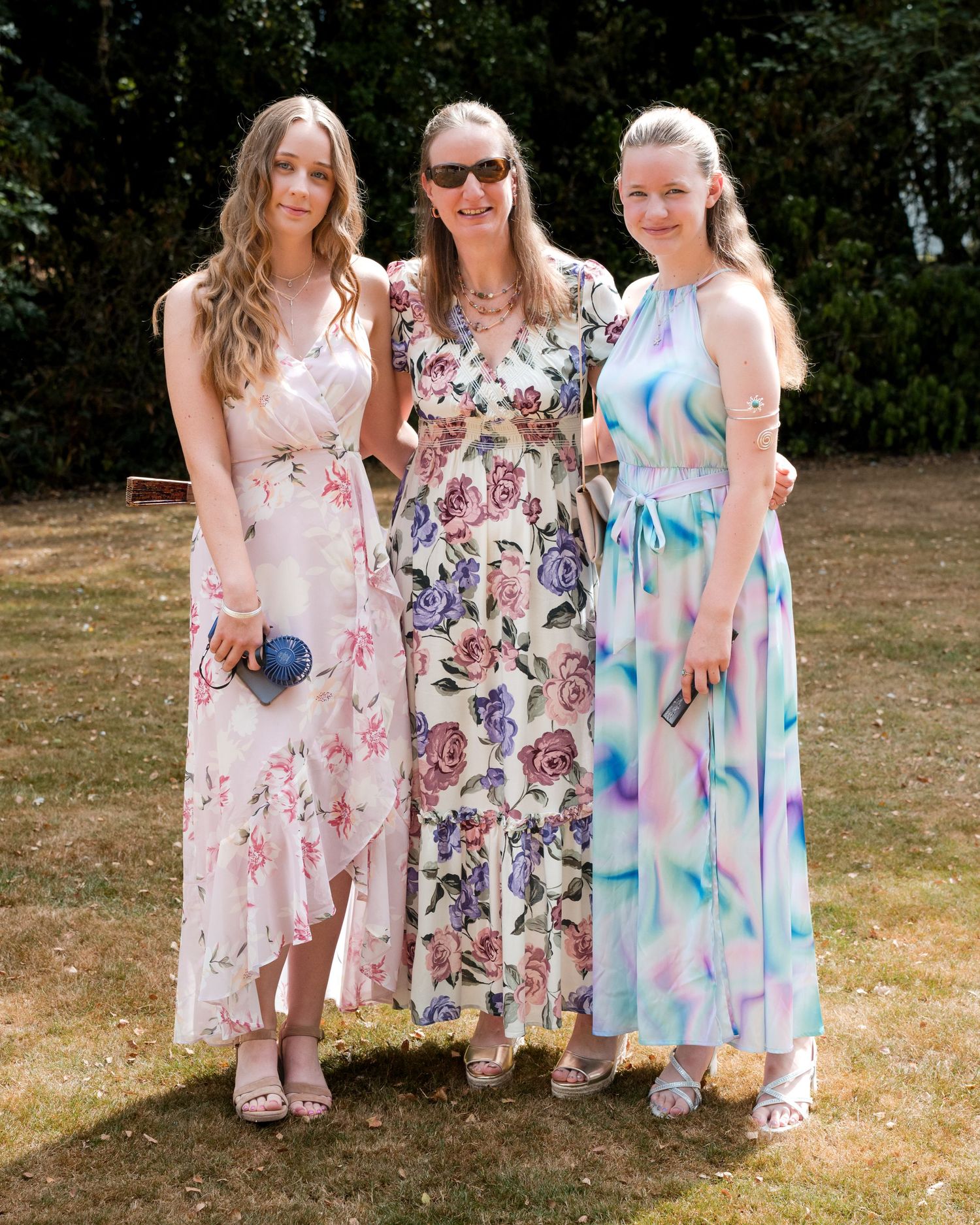Three women in floral maxi dresses posing together outdoors on a sunny day at a garden event.