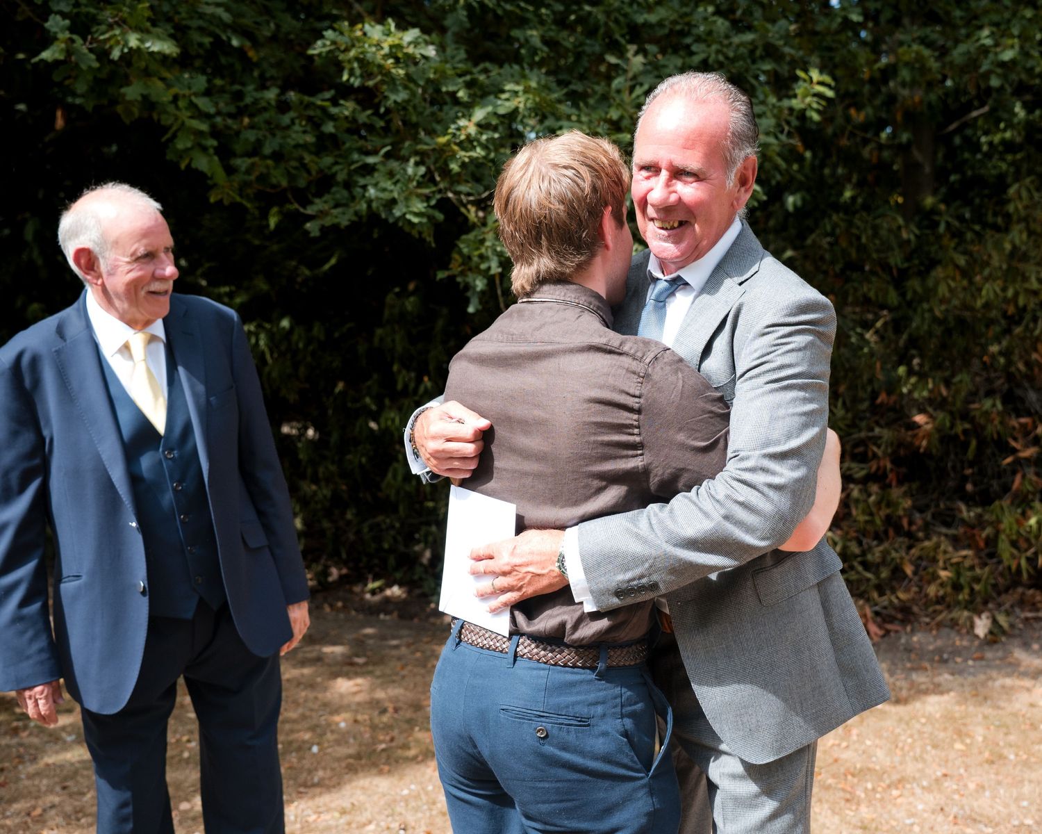 Two men share a warm hug outdoors at a formal event while an elderly man in a suit watches nearby.