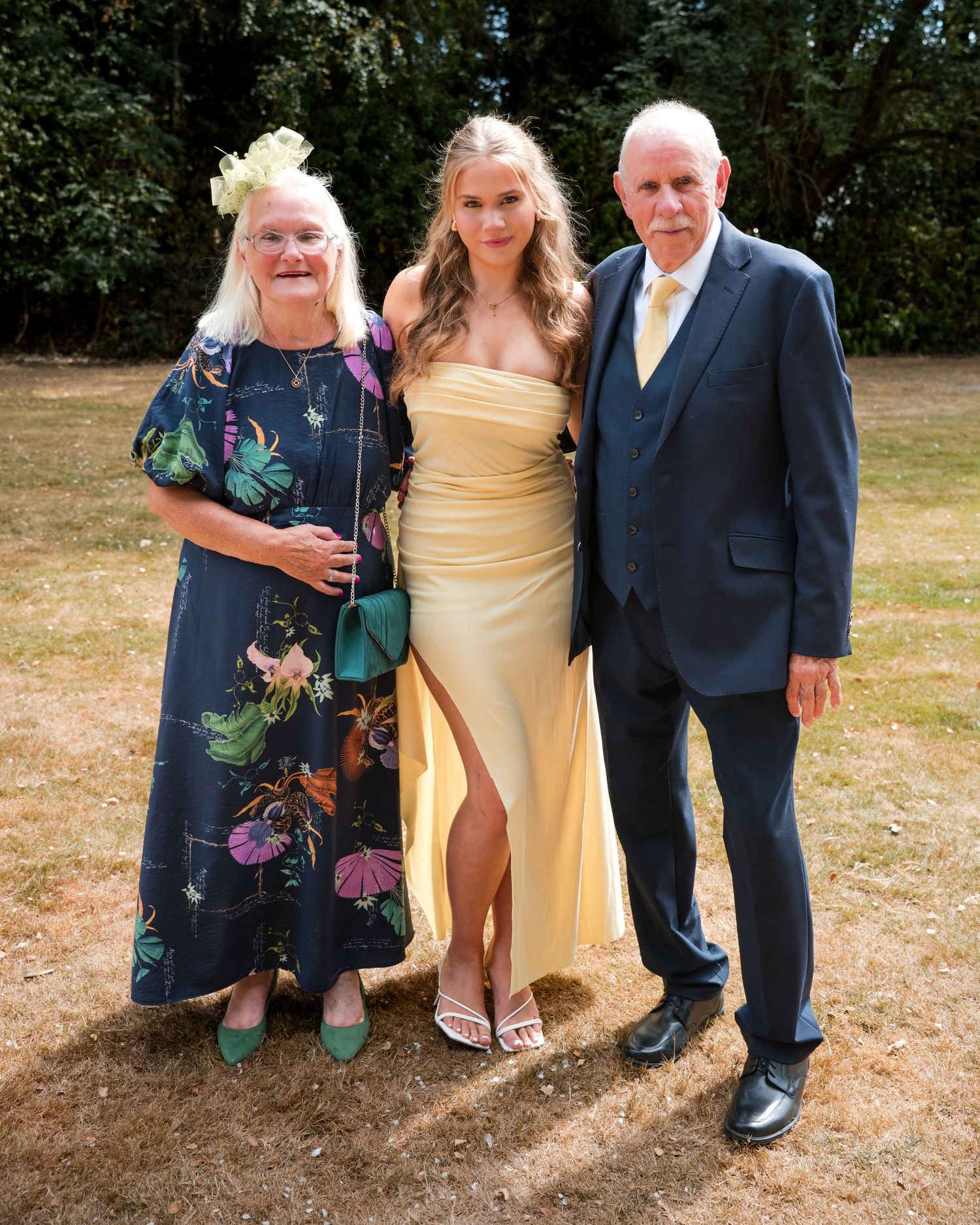 Three people posing outdoors at a wedding, wearing formal attire including a yellow dress and navy suit.