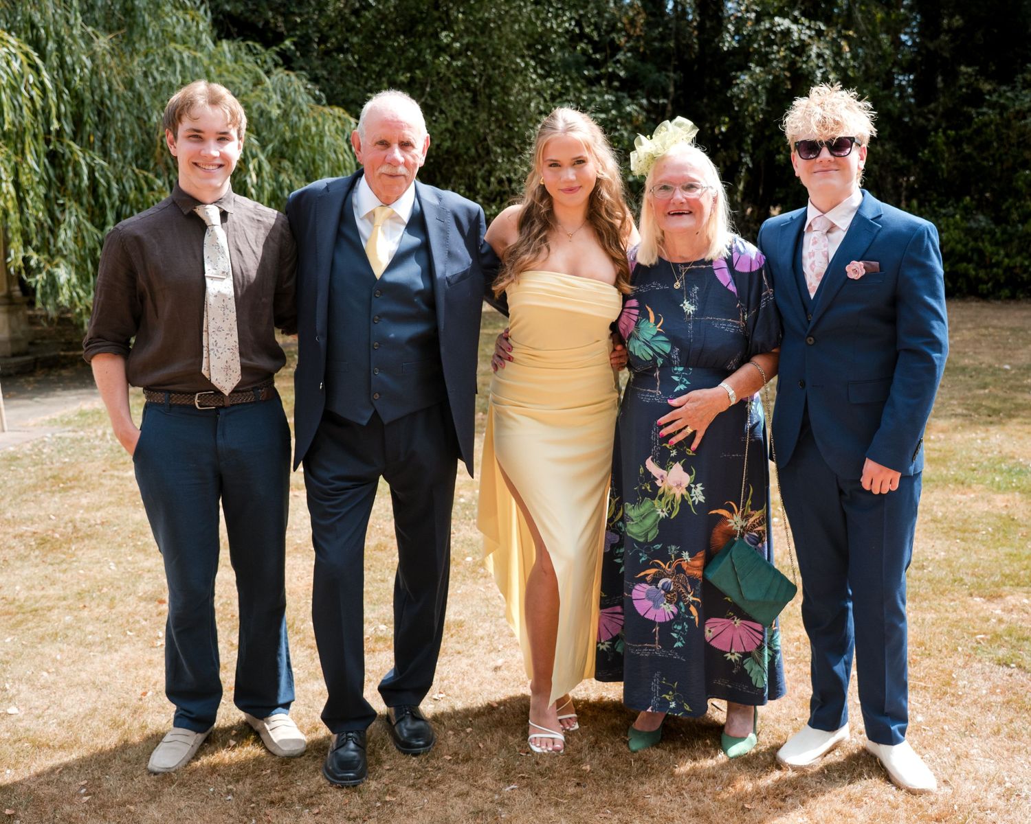 Group of five people posing outdoors at a garden event, dressed in formal and semi-formal attire on a sunny day.