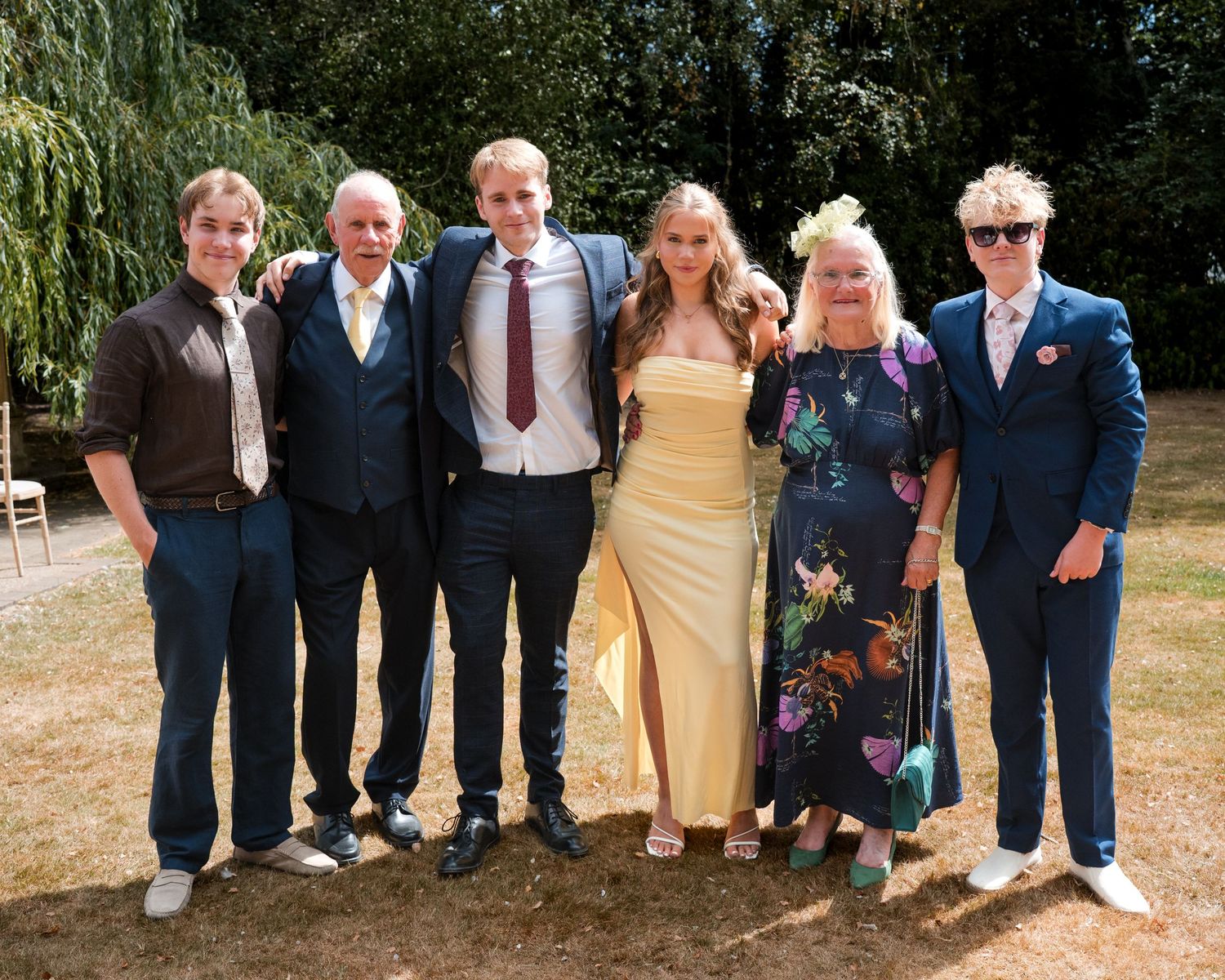 Six people dressed in formal attire pose together outdoors at a sunny wedding celebration surrounded by lush green trees.