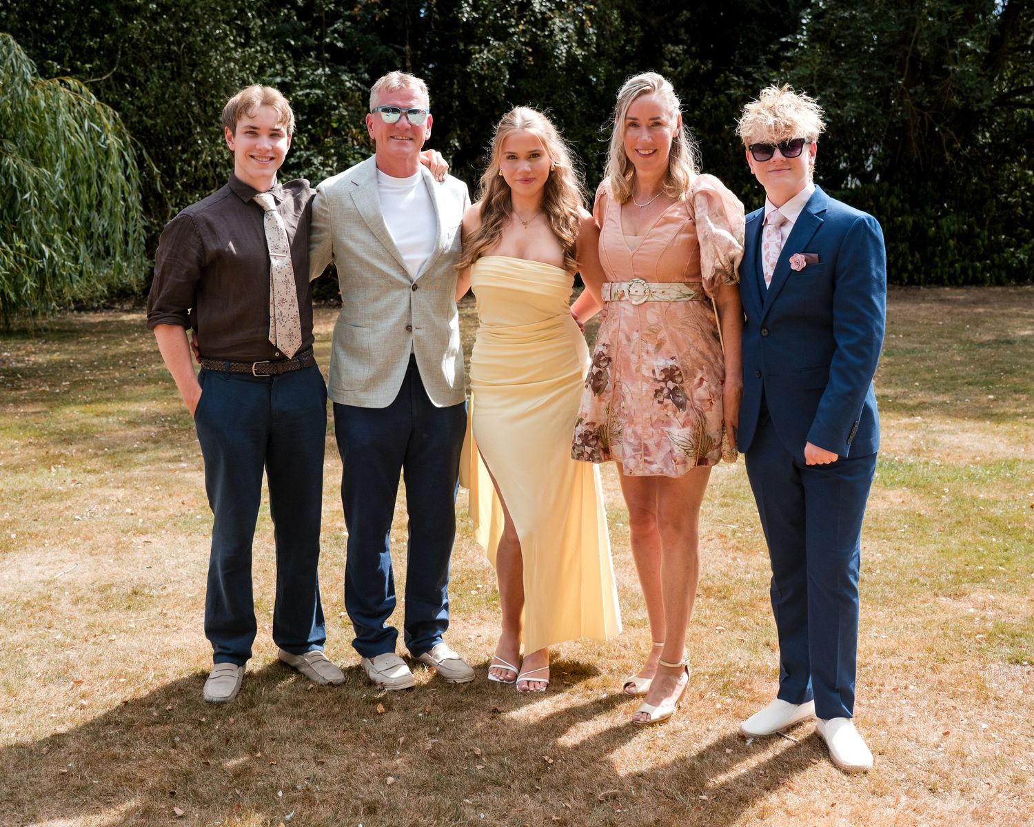 Group of five people posing outdoors in formal attire on a sunny day in a garden setting.