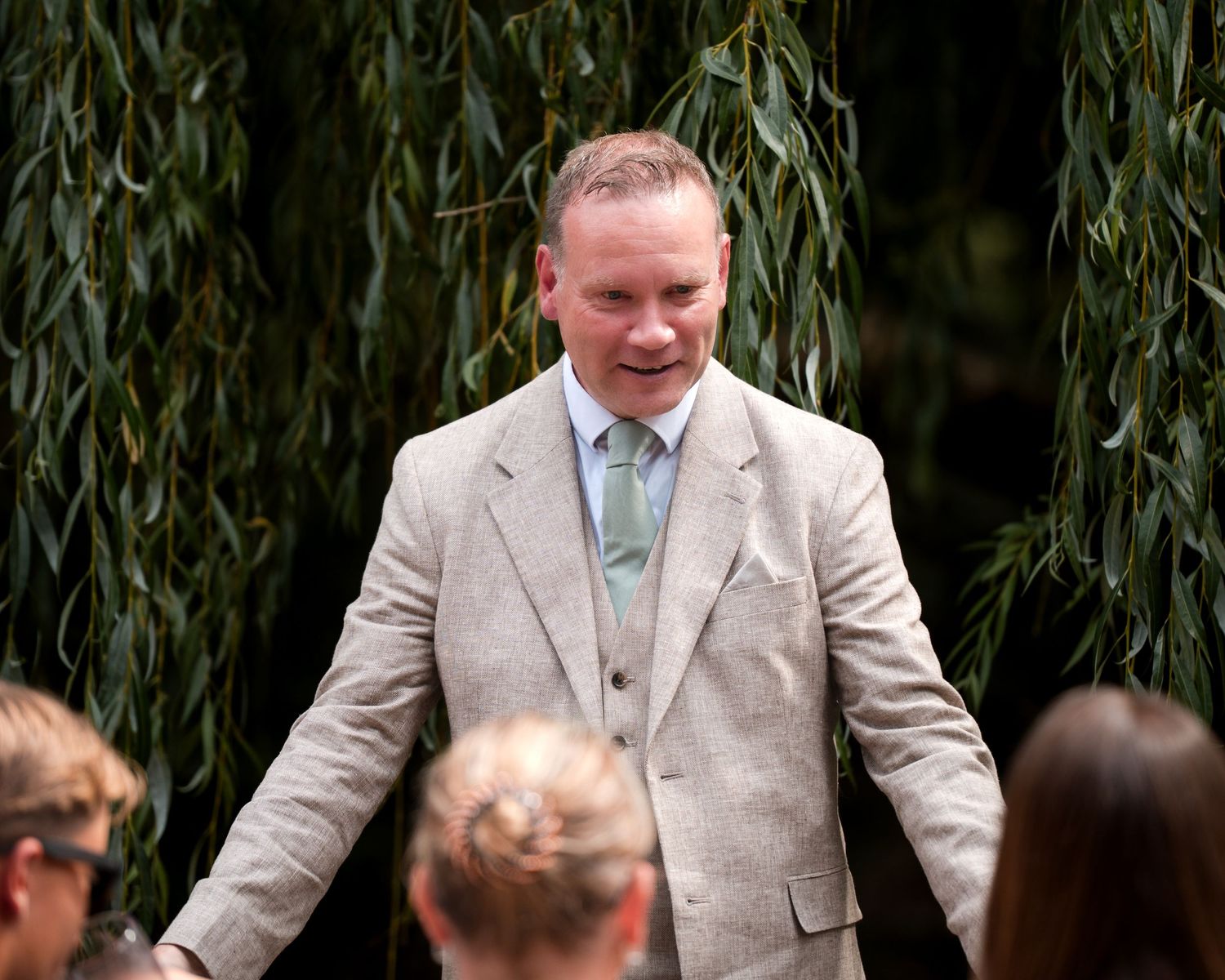 Man in beige suit with green tie smiling outdoors at a wedding ceremony surrounded by willow trees.