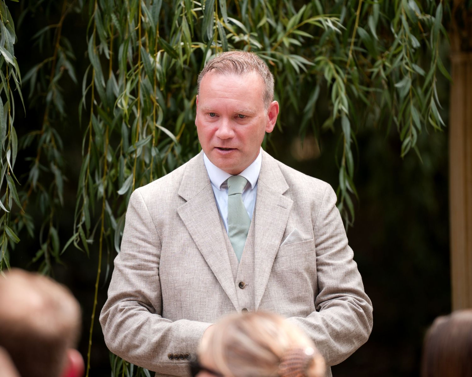 Man in beige suit with mint tie speaking outdoors under willow tree at a garden event.