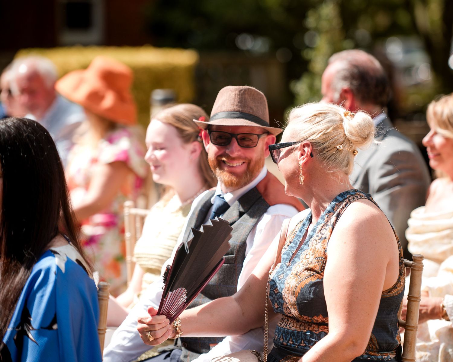 Smiling wedding guests in summer attire enjoying an outdoor ceremony, man in fedora hat and sunglasses.