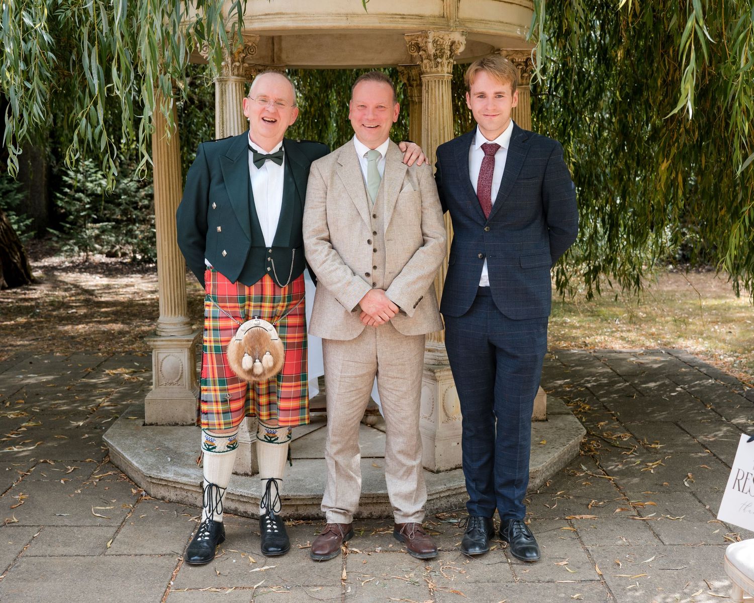 Three men posing outdoors, one wearing a traditional Scottish kilt, at a formal garden wedding celebration.