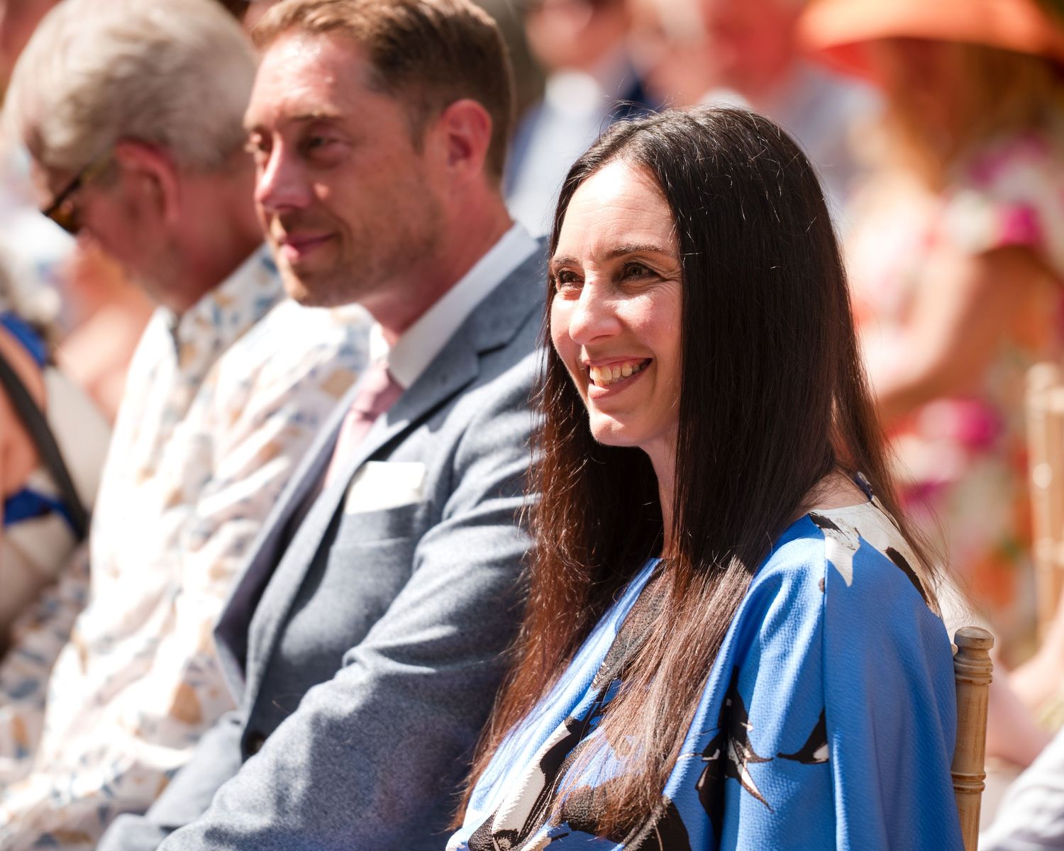 A smiling woman with long dark hair in a blue outfit seated at an outdoor wedding ceremony on a sunny day.