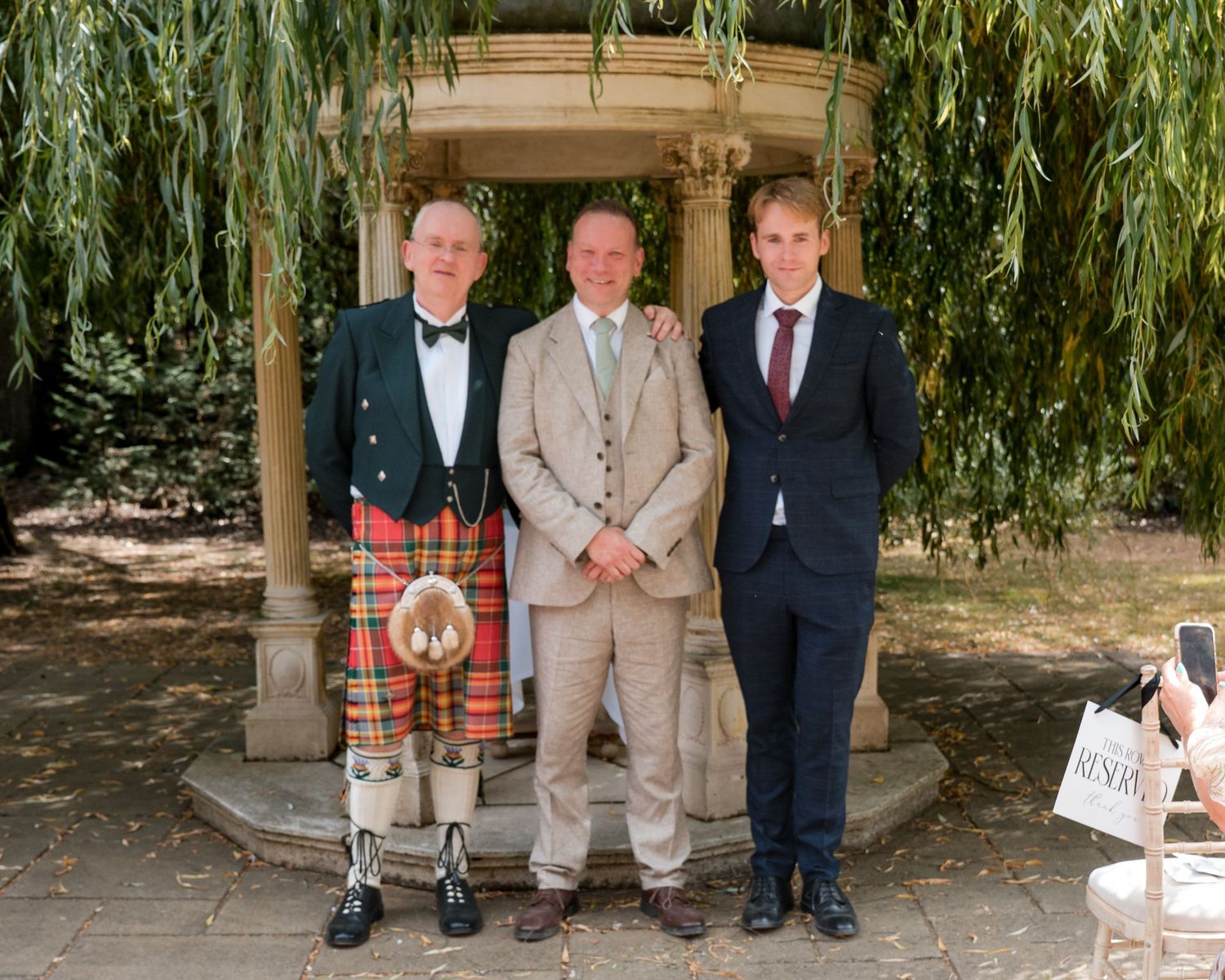 Three men posing outdoors, one wearing a Scottish kilt, at a garden wedding ceremony.