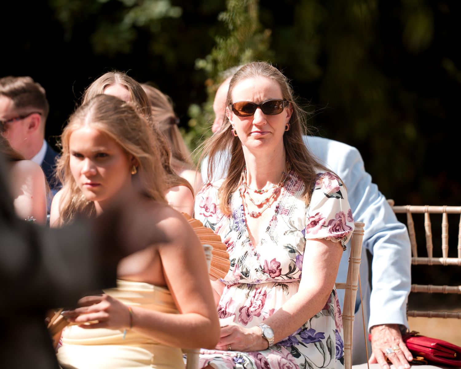 Woman in floral dress and sunglasses seated at an outdoor wedding ceremony among other guests.
