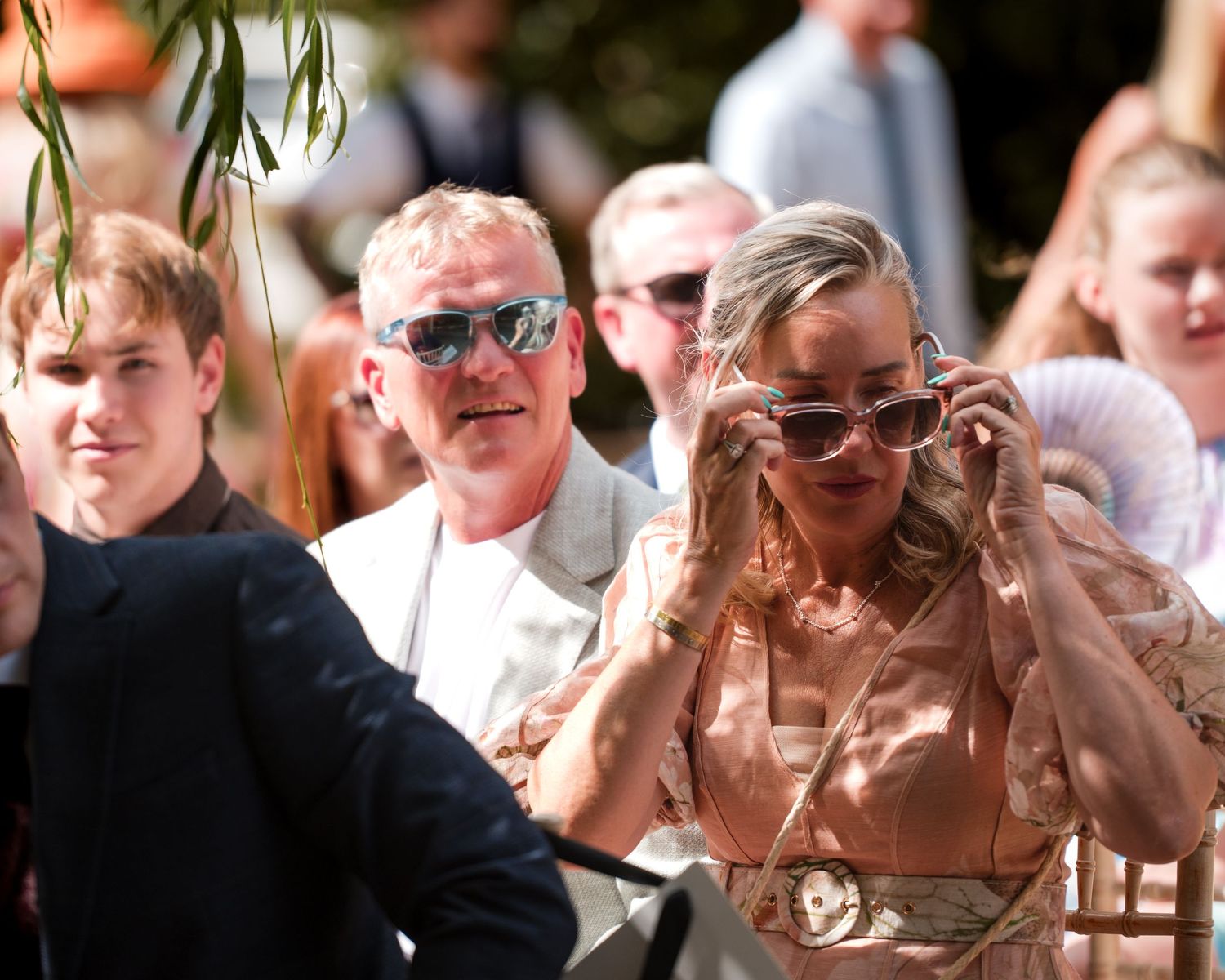 Guests at an outdoor wedding ceremony, woman adjusting sunglasses in elegant attire on a sunny day.