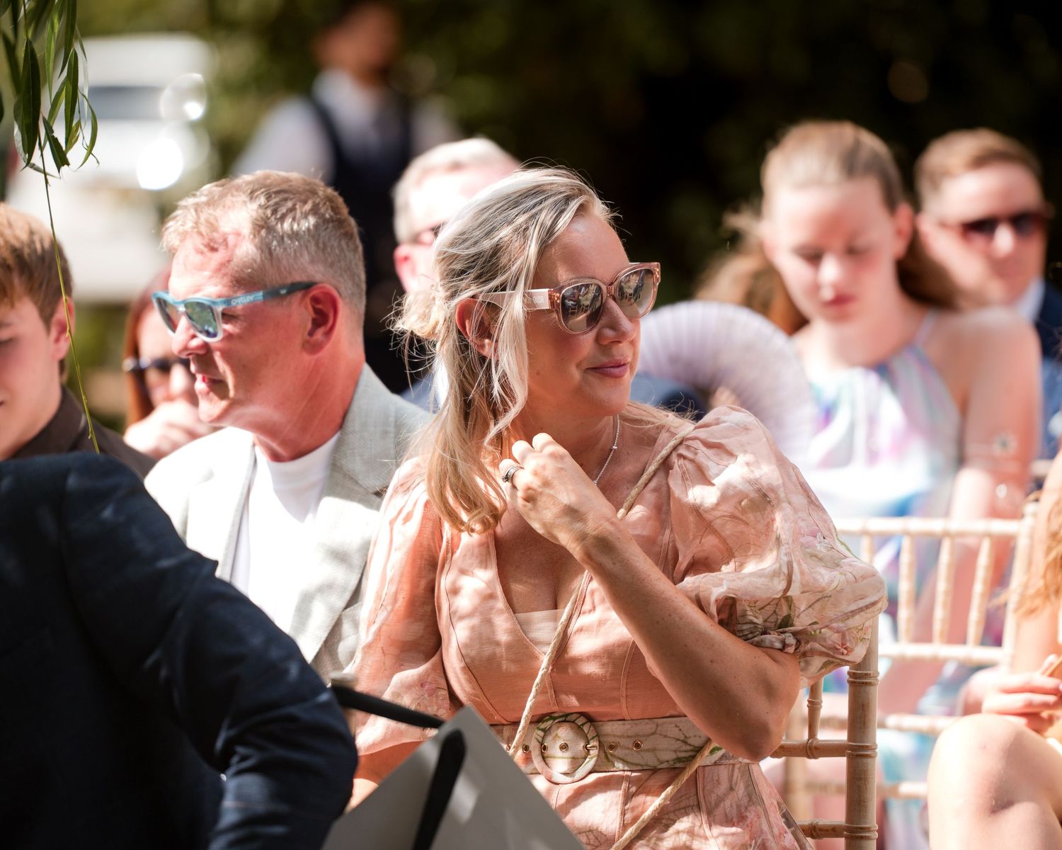 Elegantly dressed wedding guests seated outdoors, woman in floral dress wearing sunglasses enjoys sunny ceremony.