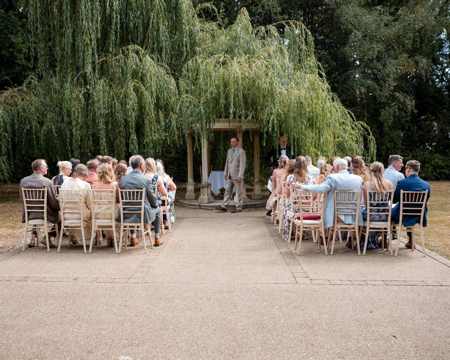 Outdoor wedding ceremony with guests seated in rows before an officiant under a weeping willow tree.