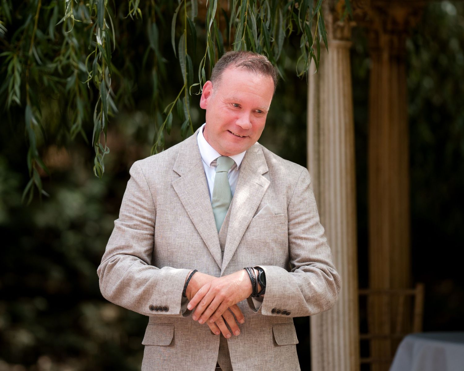 Smiling man in beige linen suit with mint tie standing outdoors under weeping willow tree.