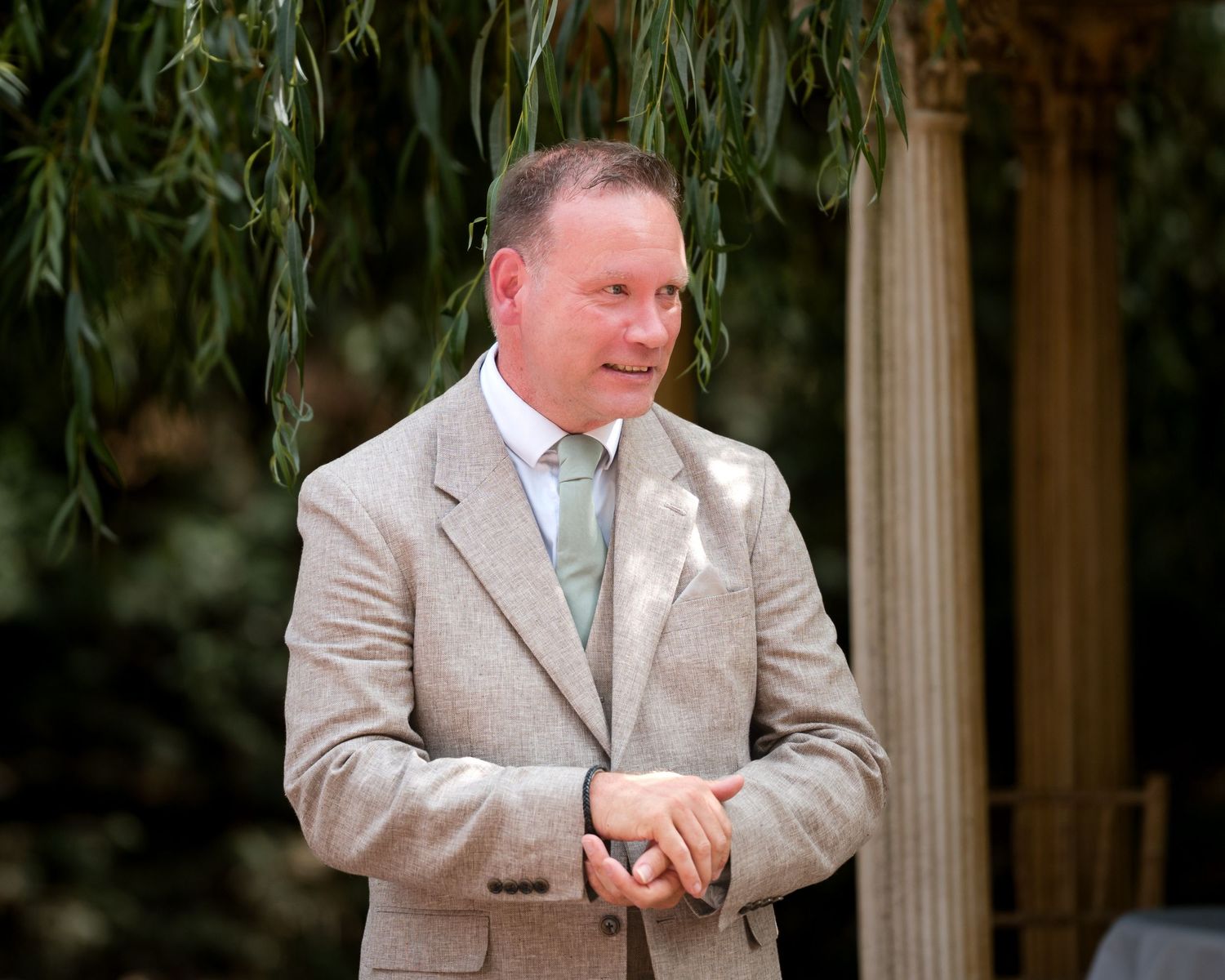 Elegant man in beige suit with mint tie stands outdoors among lush green weeping willow trees.