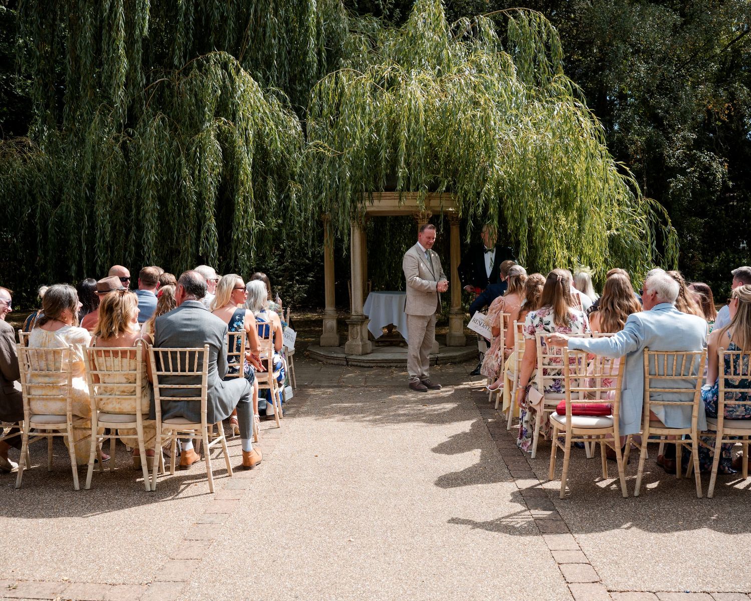 Outdoor wedding ceremony with guests seated on wooden chairs beneath a weeping willow tree in a sunny garden.