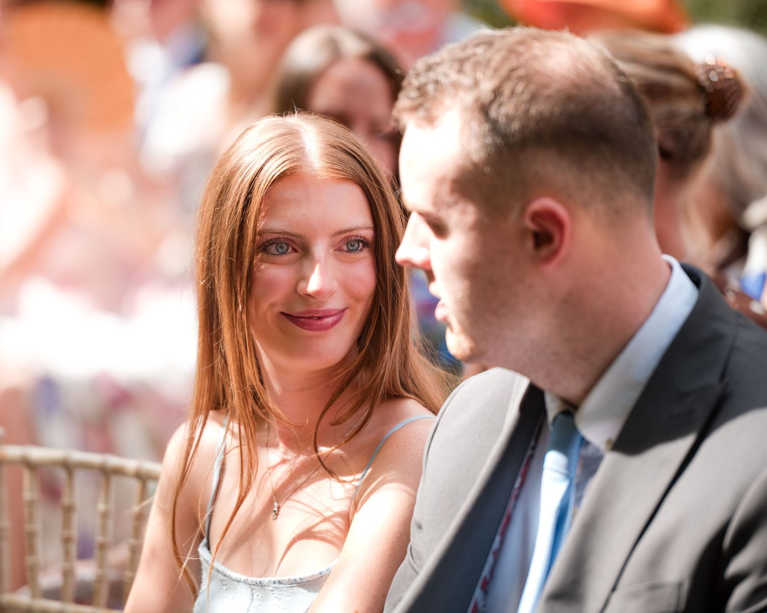 A red-haired woman in a white dress smiles at a man in a gray suit at an outdoor wedding ceremony.