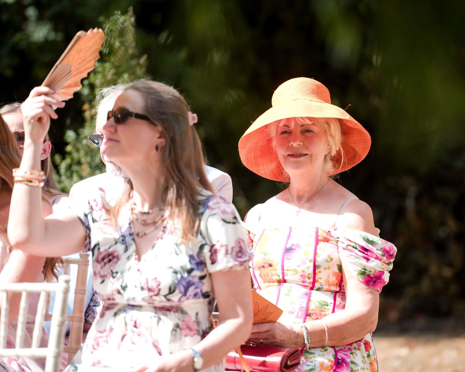 Two elegantly dressed women at an outdoor summer event, one wearing an orange hat and floral dress, fanning themselves in the heat.