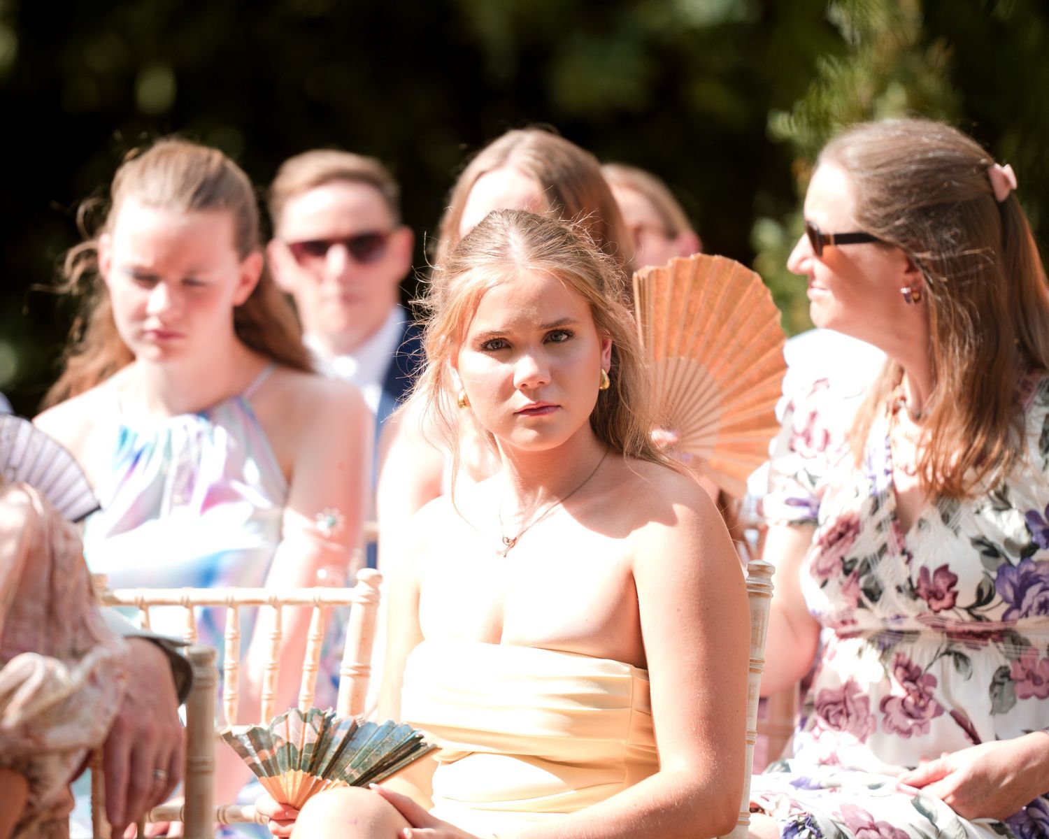 Women in summer dresses sitting outdoors at a garden event, holding decorative fans on a sunny day.