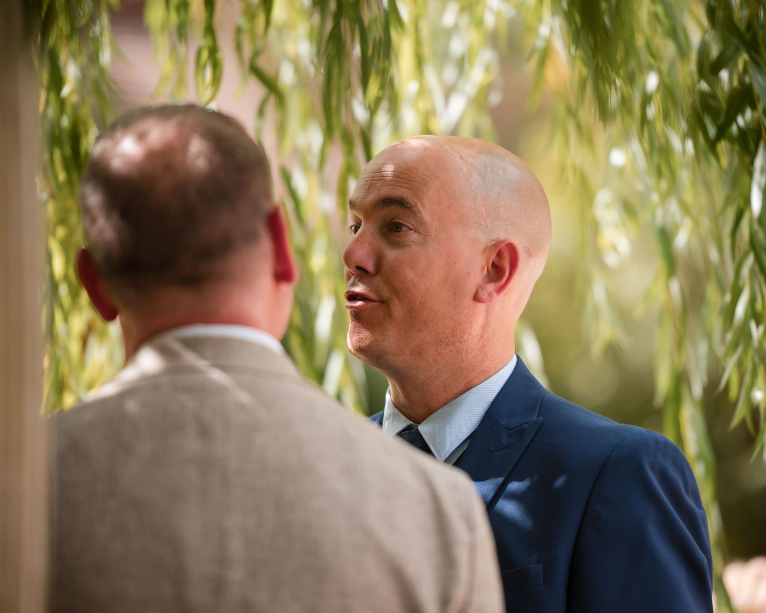 Two men in suits conversing outdoors under weeping willow trees at a formal event.