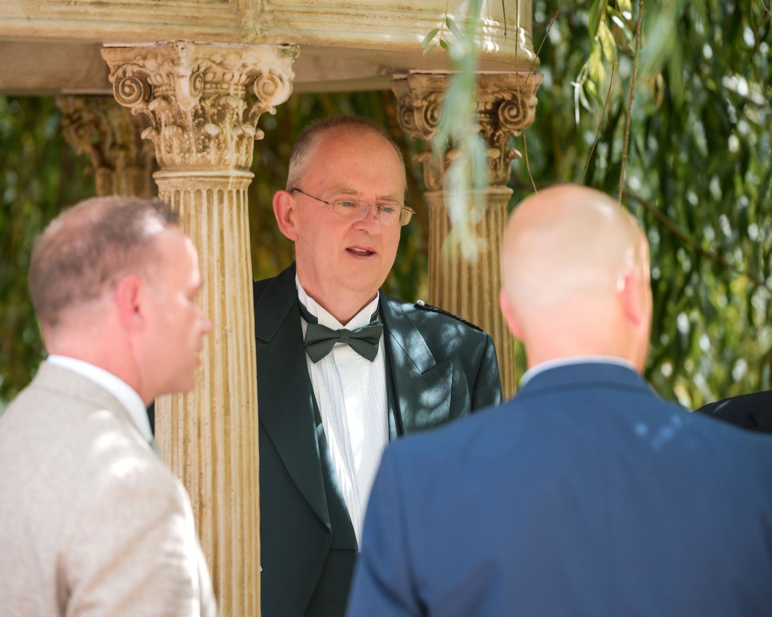 Man in tuxedo with bow tie speaks with guests under ornate stone gazebo columns at outdoor wedding ceremony.