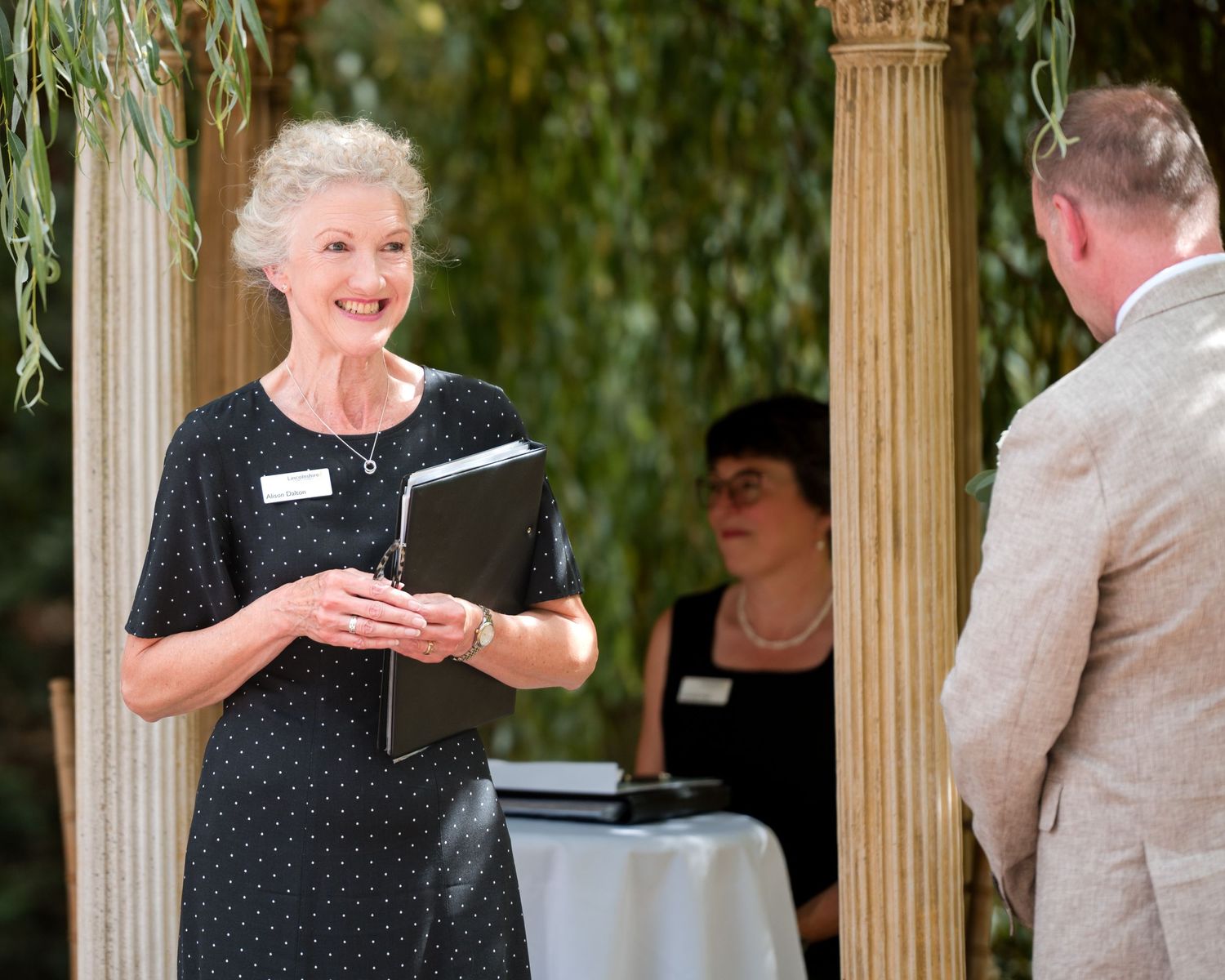 Elegant woman in black polka dot dress holding a binder, smiling at an outdoor garden ceremony event.