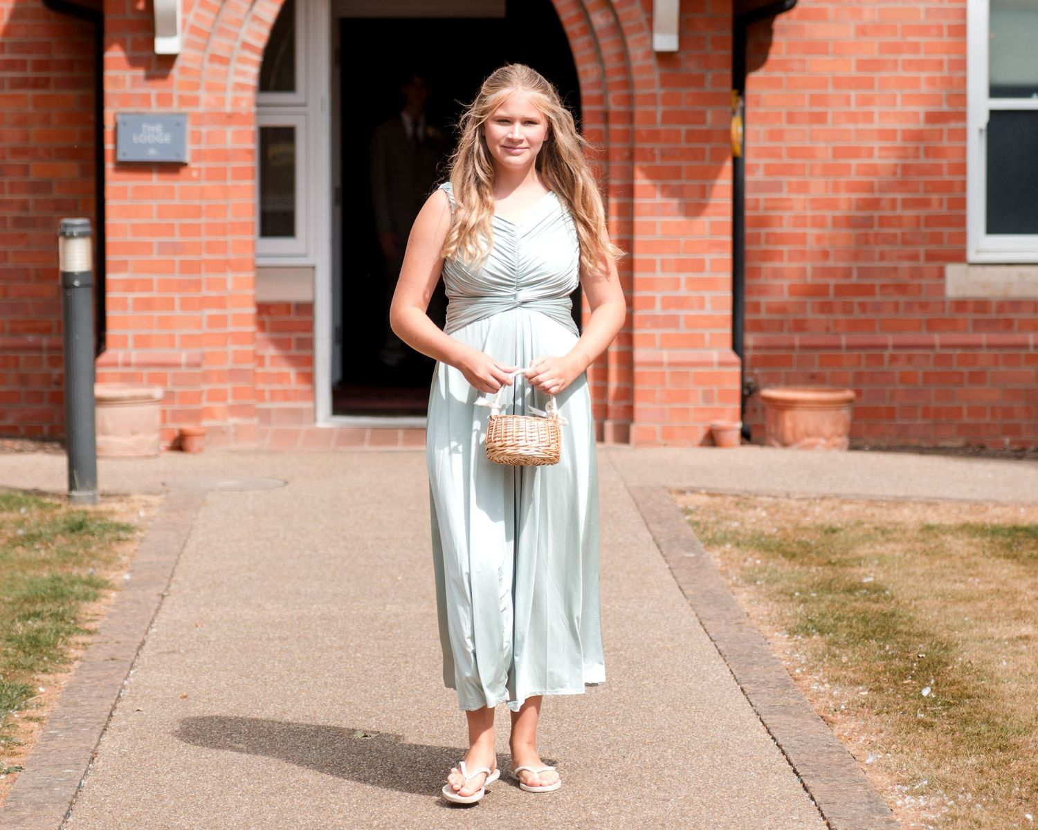 Young woman in mint green maxi dress holding wicker bag, standing on pathway outside red brick building.