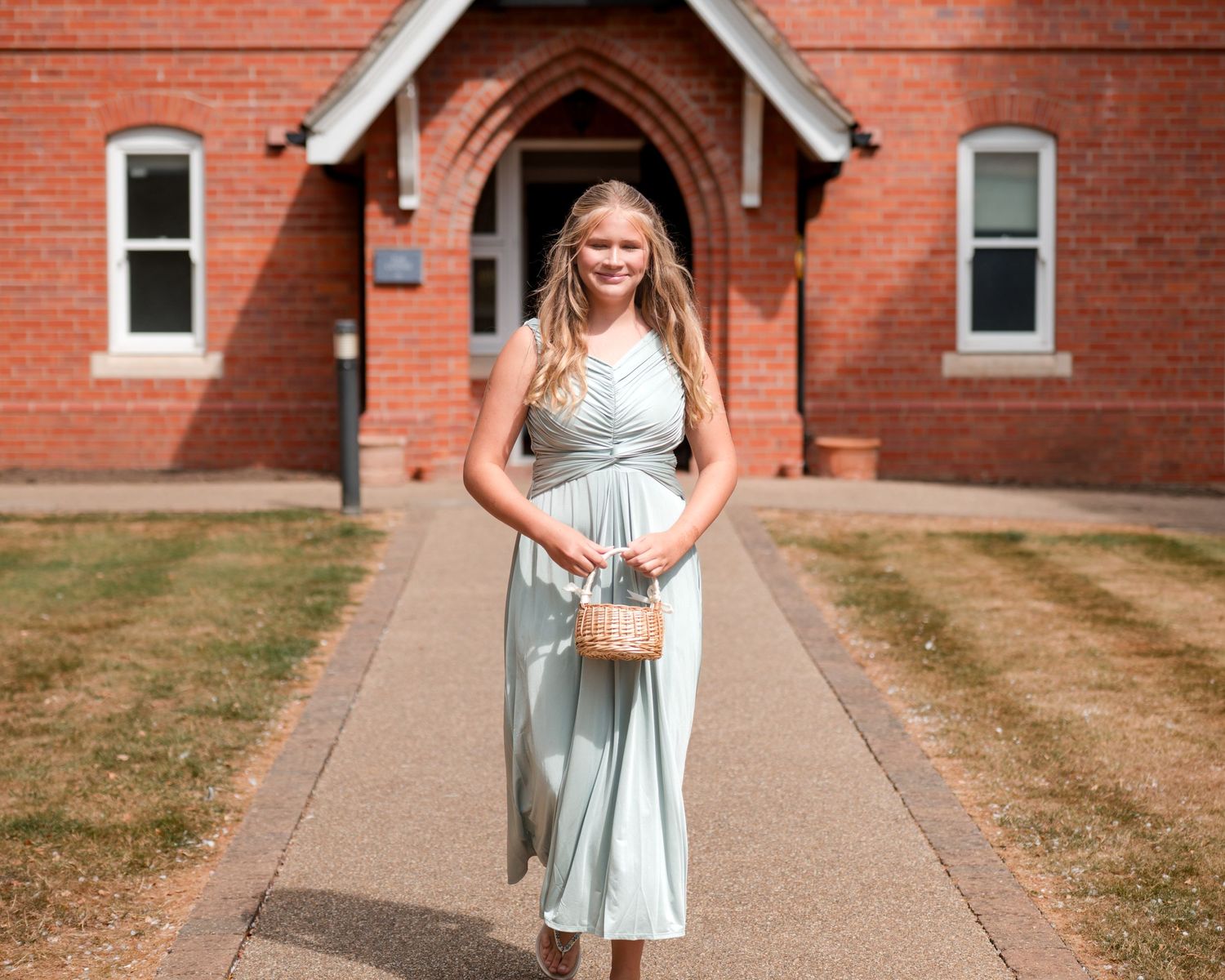 Young girl in mint green dress holding wicker purse walks along church pathway on sunny day.