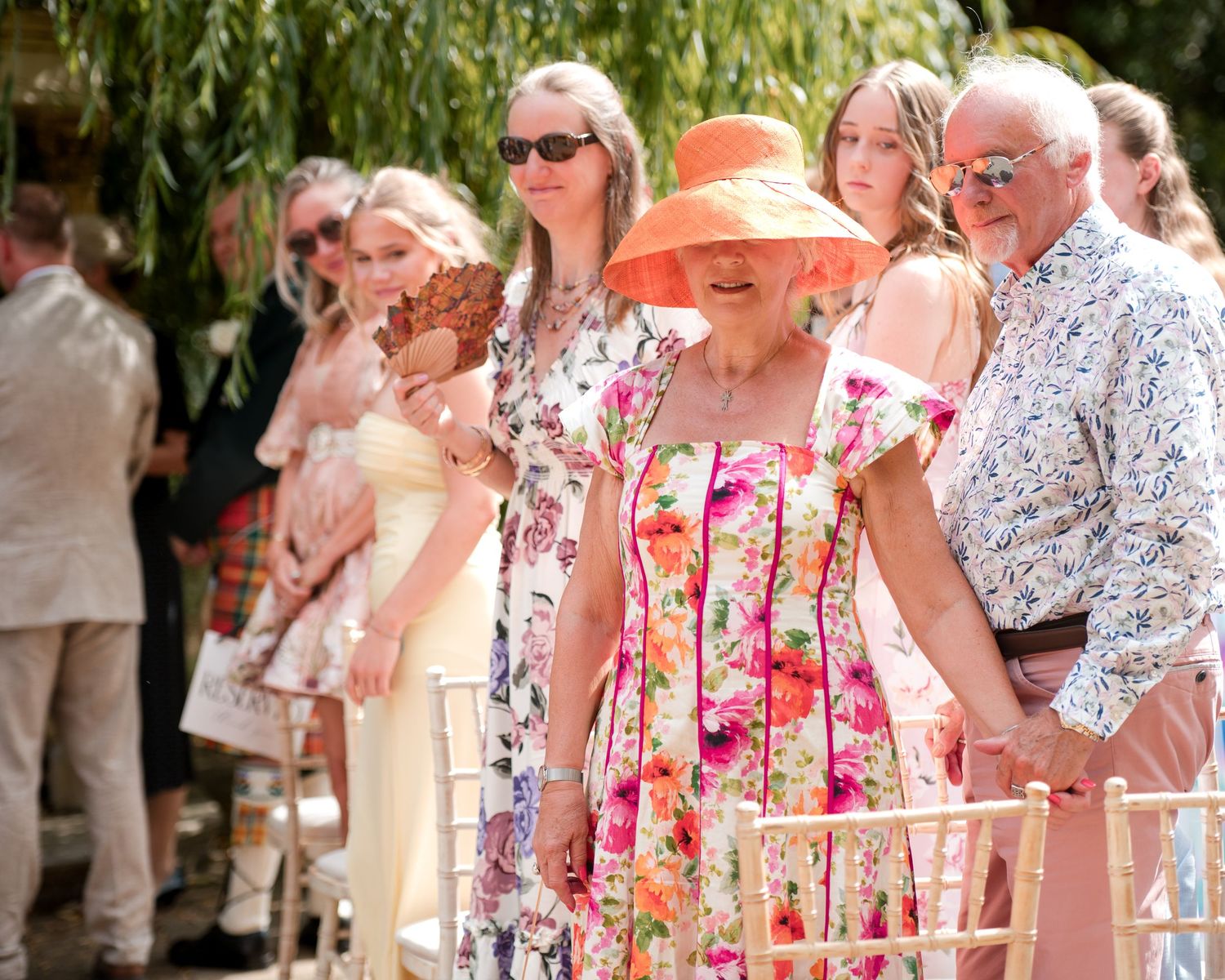 Elderly woman in floral dress and orange hat walks with man at outdoor wedding ceremony.